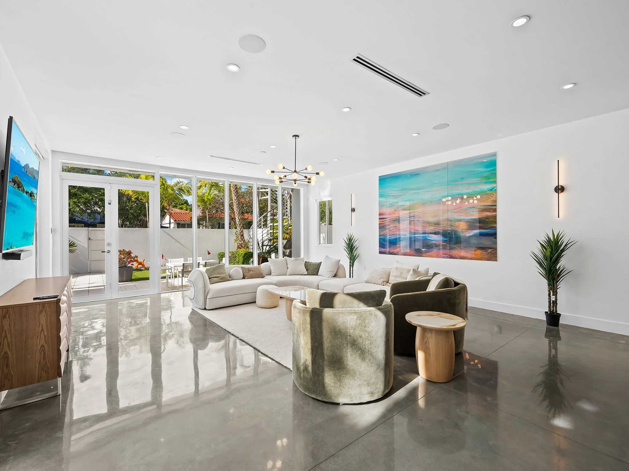 A modern living room with white walls, a large abstract painting, a white sectional sofa, and a mix of wood and velvet chairs. Large windows and glass doors let in natural light, showing a backyard with trees, a patio table, and outdoor seating. The 