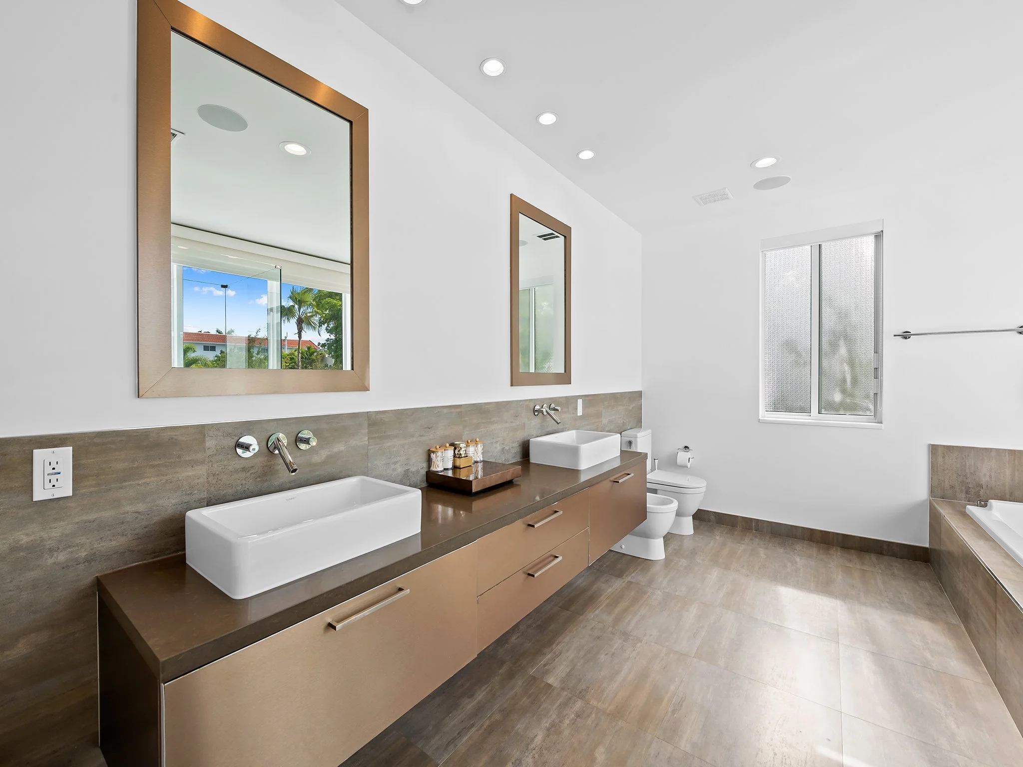 Modern bathroom with two rectangular vessel sinks on a wooden vanity with drawers, large mirrors, a window, and a toilet.