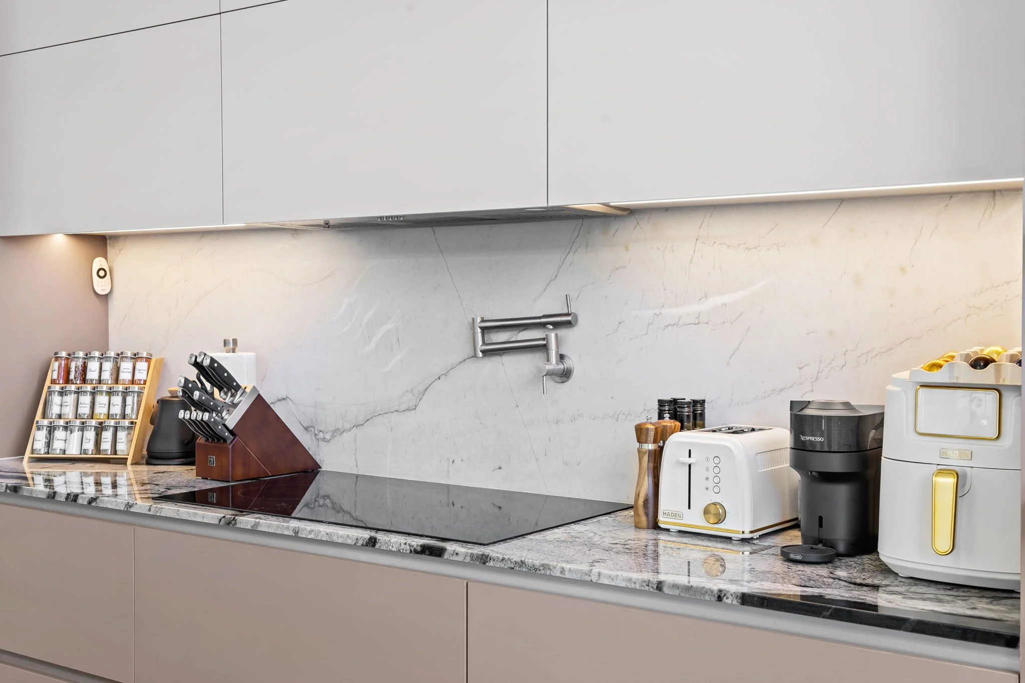 Kitchen countertop with a spice rack, knives in a wooden holder, small appliances including a toaster, coffee machine, and kettle, with a marble backsplash and upper cabinets.