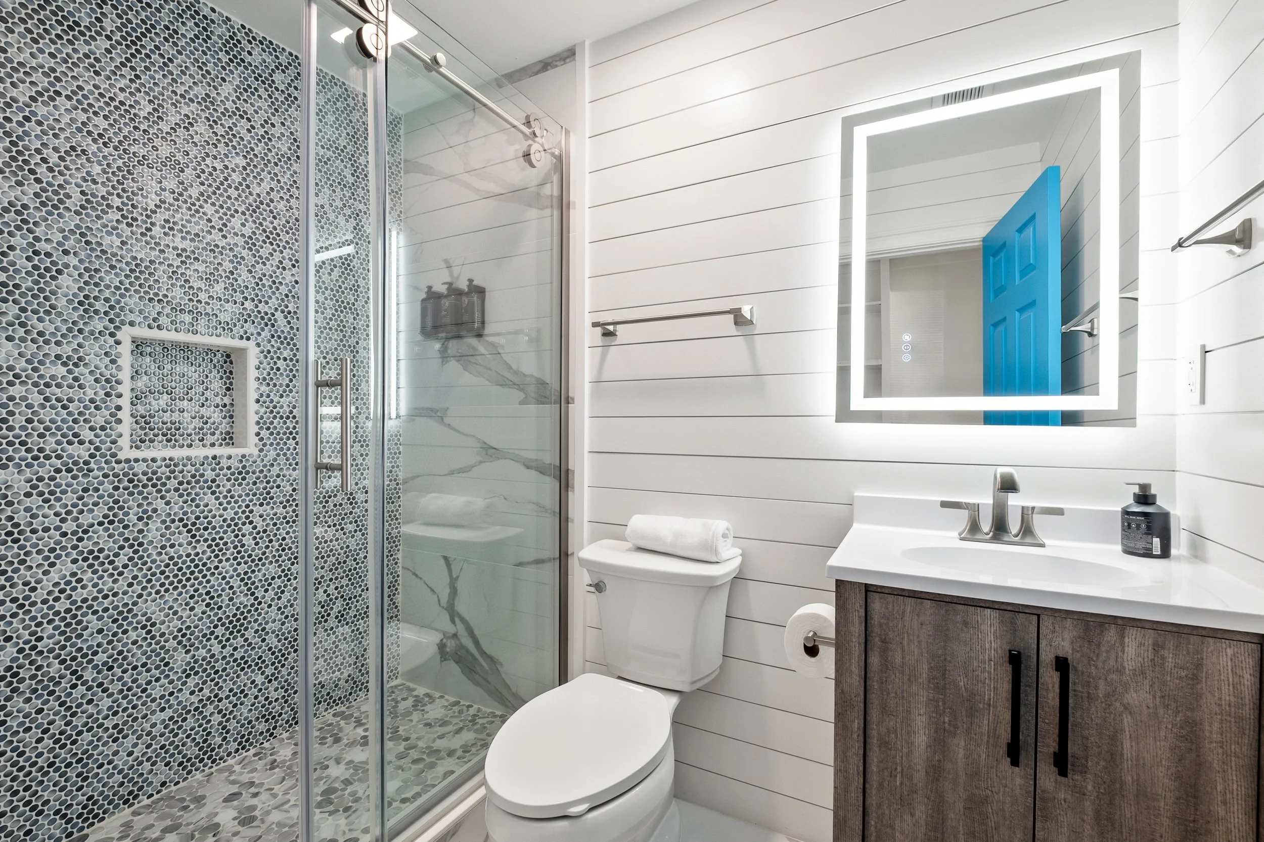 Modern bathroom with a shower, toilet, and vanity. The shower has a glass door and black and white mosaic tiles. The walls are white horizontal shiplap, and there is a square mirror with LED lighting above the vanity. A blue door is partially open, r
