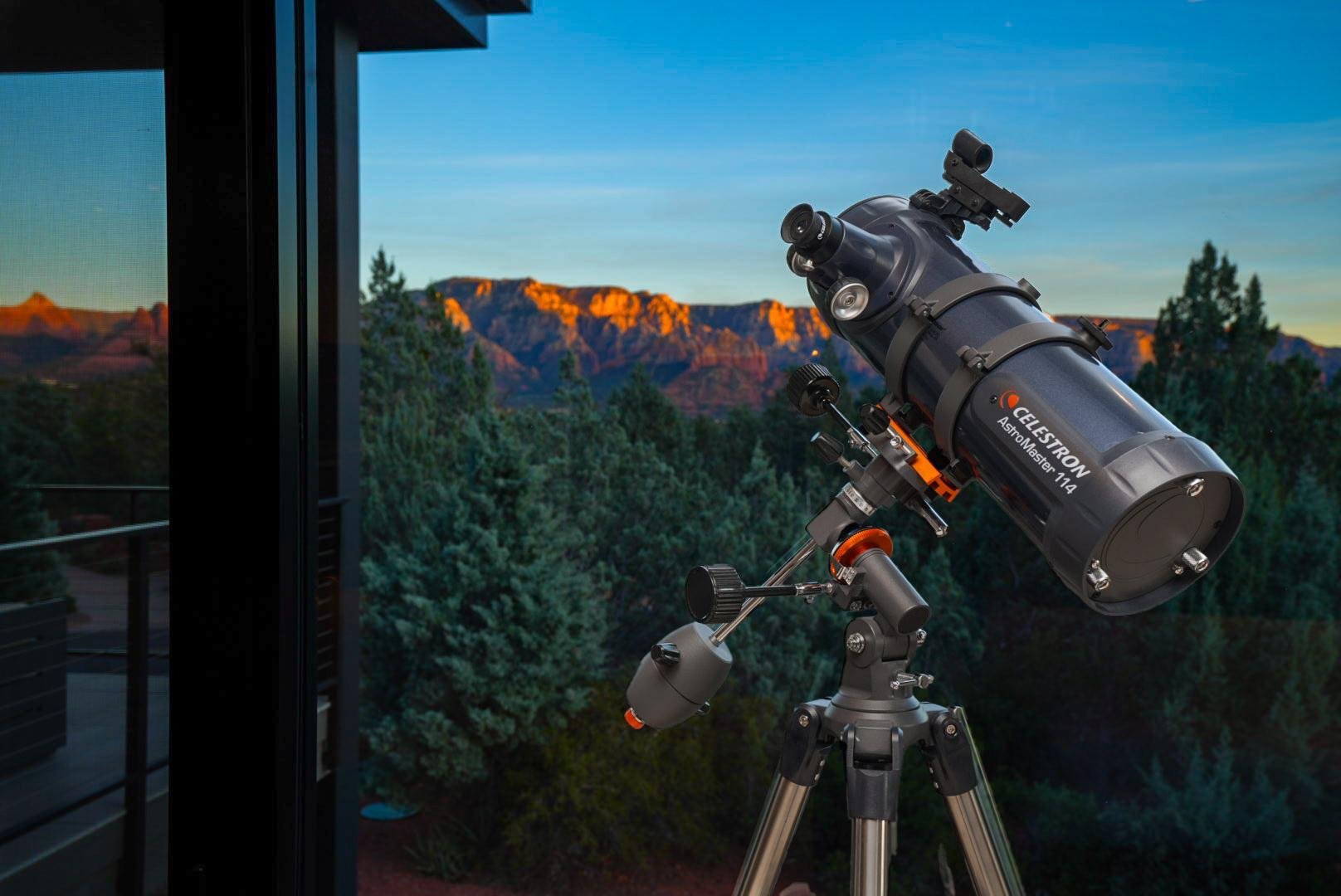 A telescope set up on a balcony with a view of distant mountains and trees at sunset.