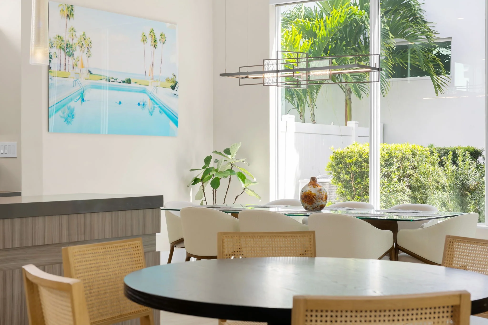 Modern dining area with white chairs around a circular black table, a large window with greenery outside, a decorative hanging light fixture, a potted plant, a butter ceramic vase, and a painting of palm trees near a pool on the wall.