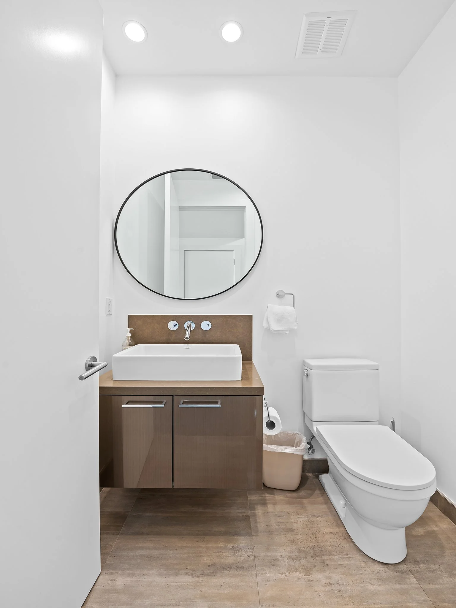 Modern bathroom with white walls and ceiling, round mirror above a white rectangular vessel sink on a wooden cabinet, a toilet, a towel on a wall hook, a trash bin, and a wall-mounted soap dispenser.