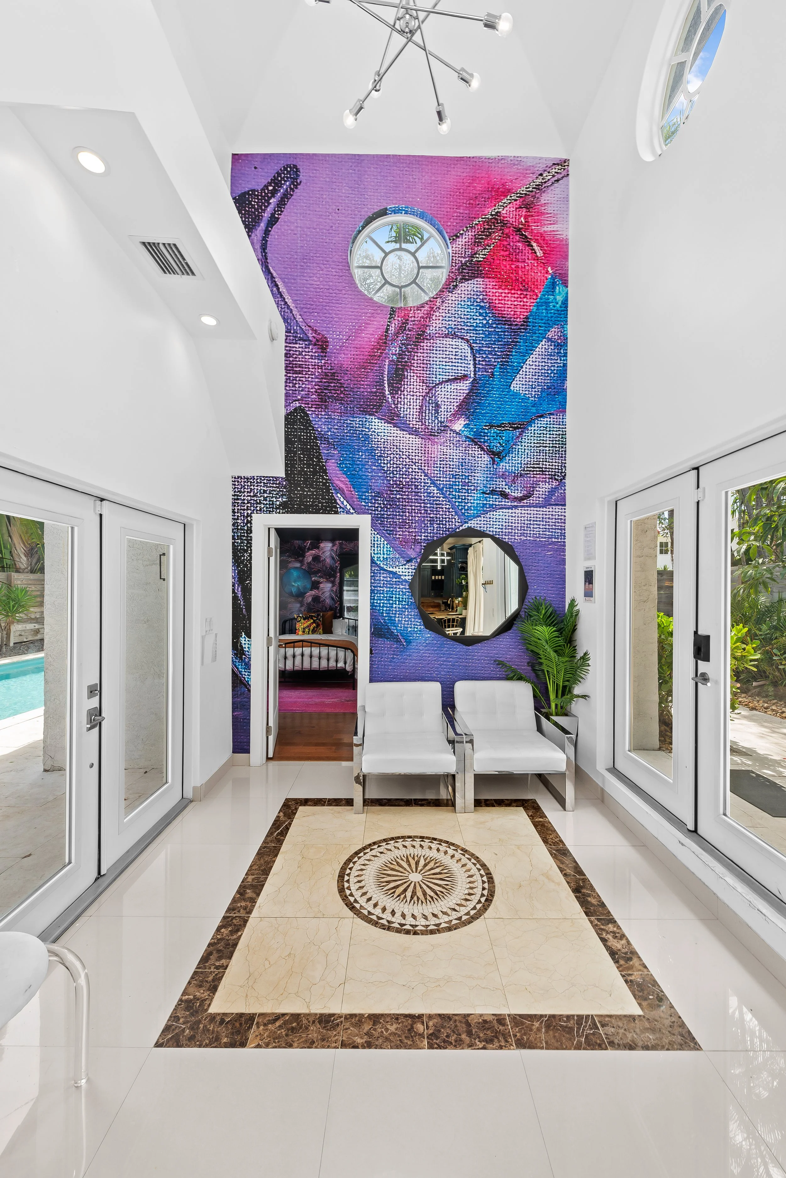 Bright interior space with white walls and a decorative tile floor, white chairs, large glass doors, a colorful mosaic mural on the wall featuring abstract art and colors like pink, blue, purple, a round window, and a view of a backyard with greenery.