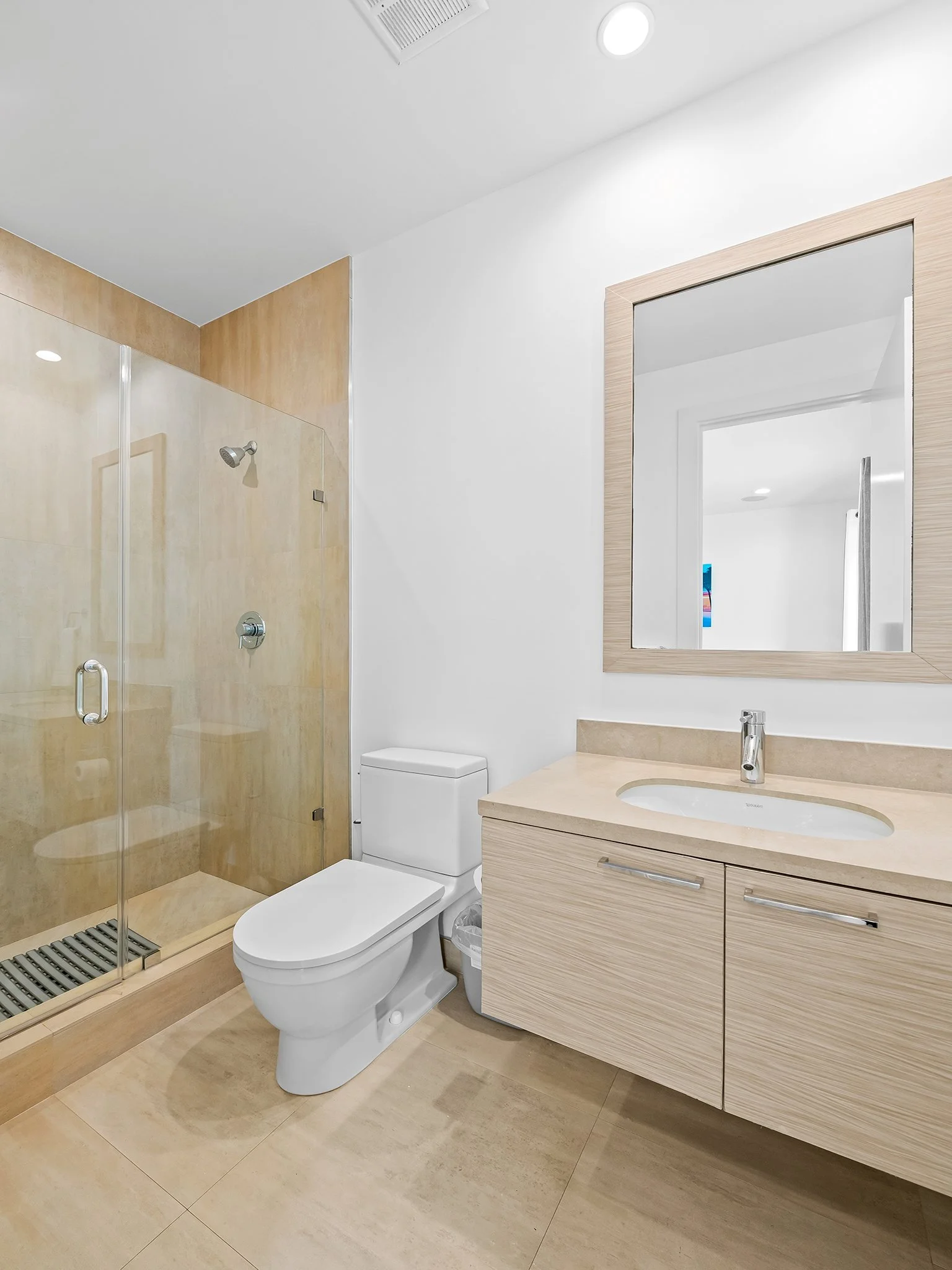 Modern bathroom with a glass-enclosed shower, a white toilet, and a wooden vanity with a sink, large mirror, and light wood finishes.