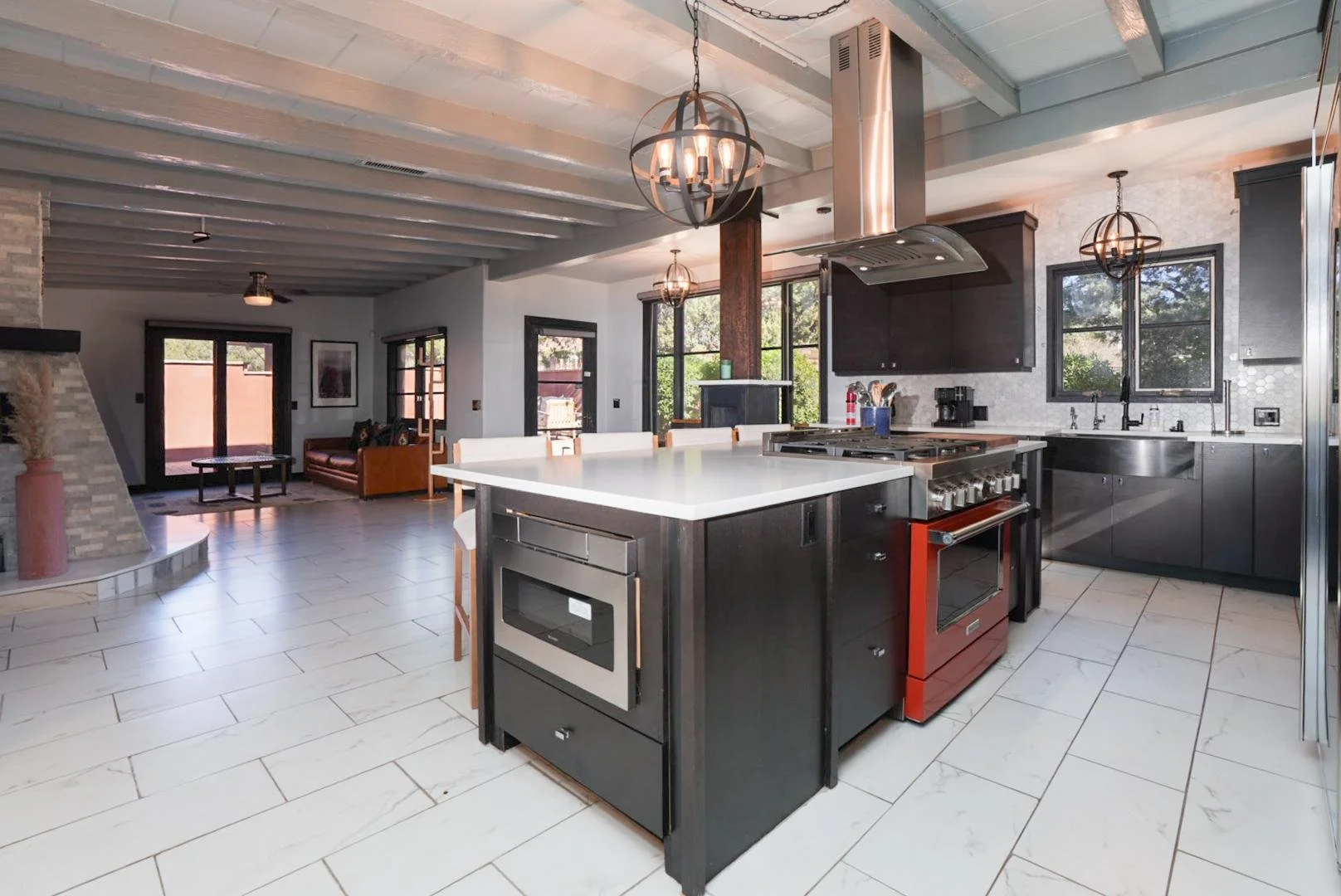 Modern open-concept kitchen with a white island, stainless steel appliances including a red oven, dark cabinetry, and black-framed windows, adjacent to a living area with a brown sofa and a fireplace.