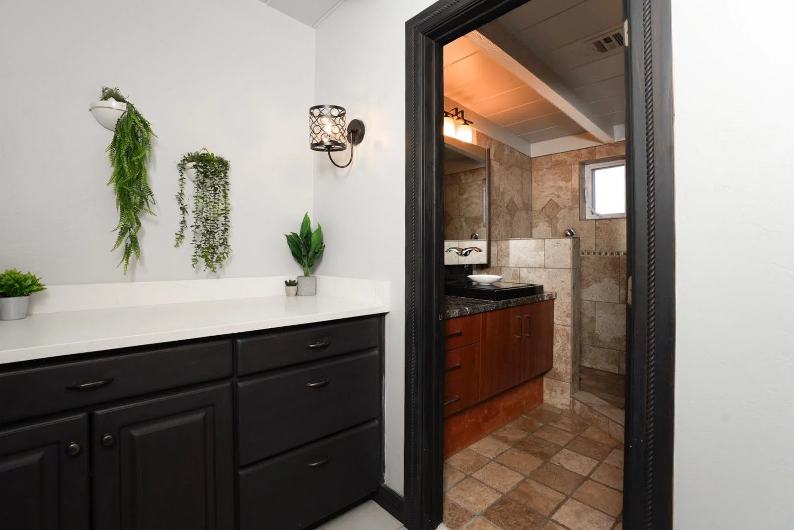 A bathroom with a black vanity and mirror, visible through a doorway. The bathroom has beige tiled walls, a small window, and a black countertop with a vessel sink.