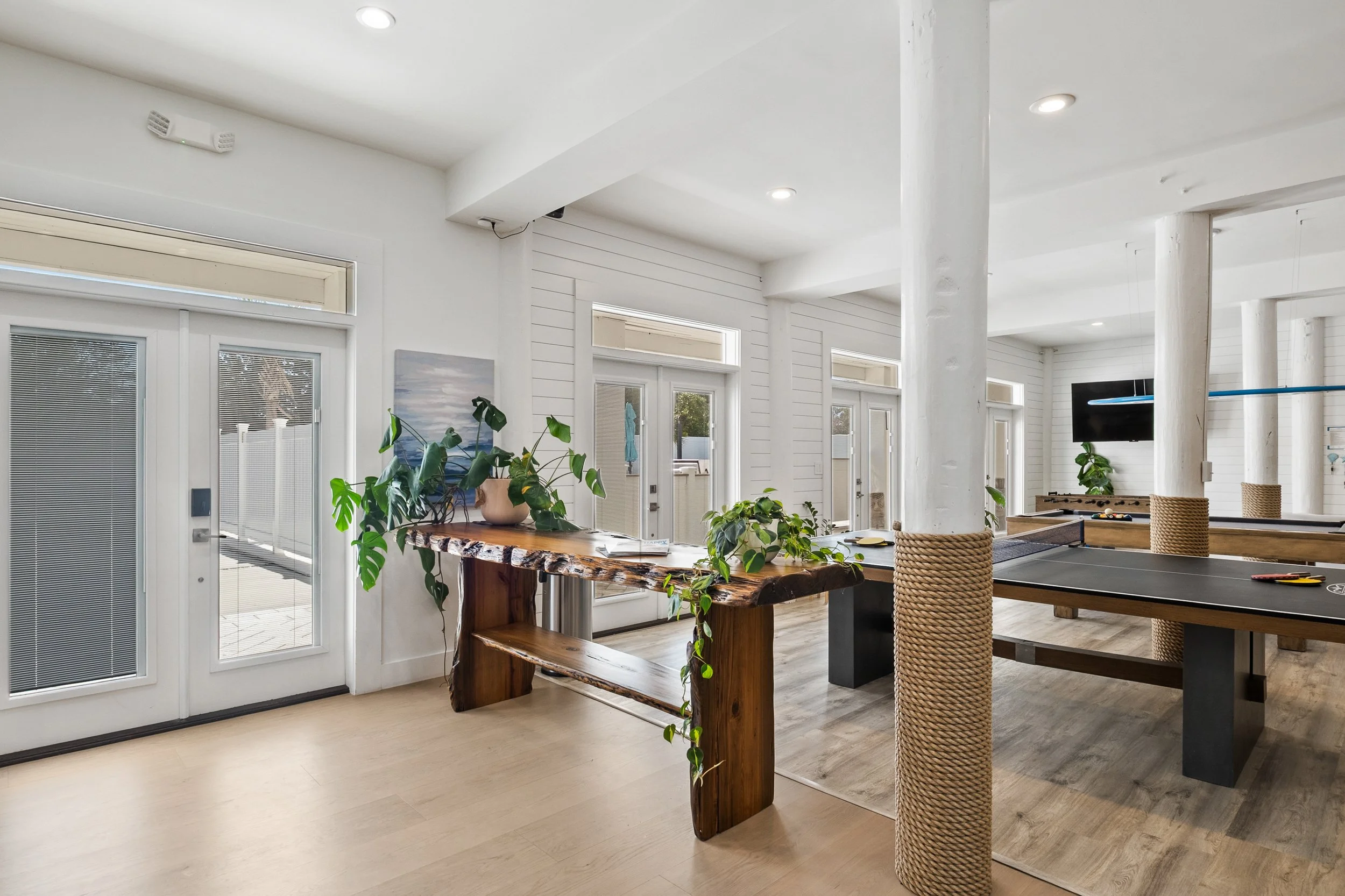 Indoor recreational room with a ping pong table, large windows, potted plants, a wooden table with plants and a notebook, and a television mounted on the wall.