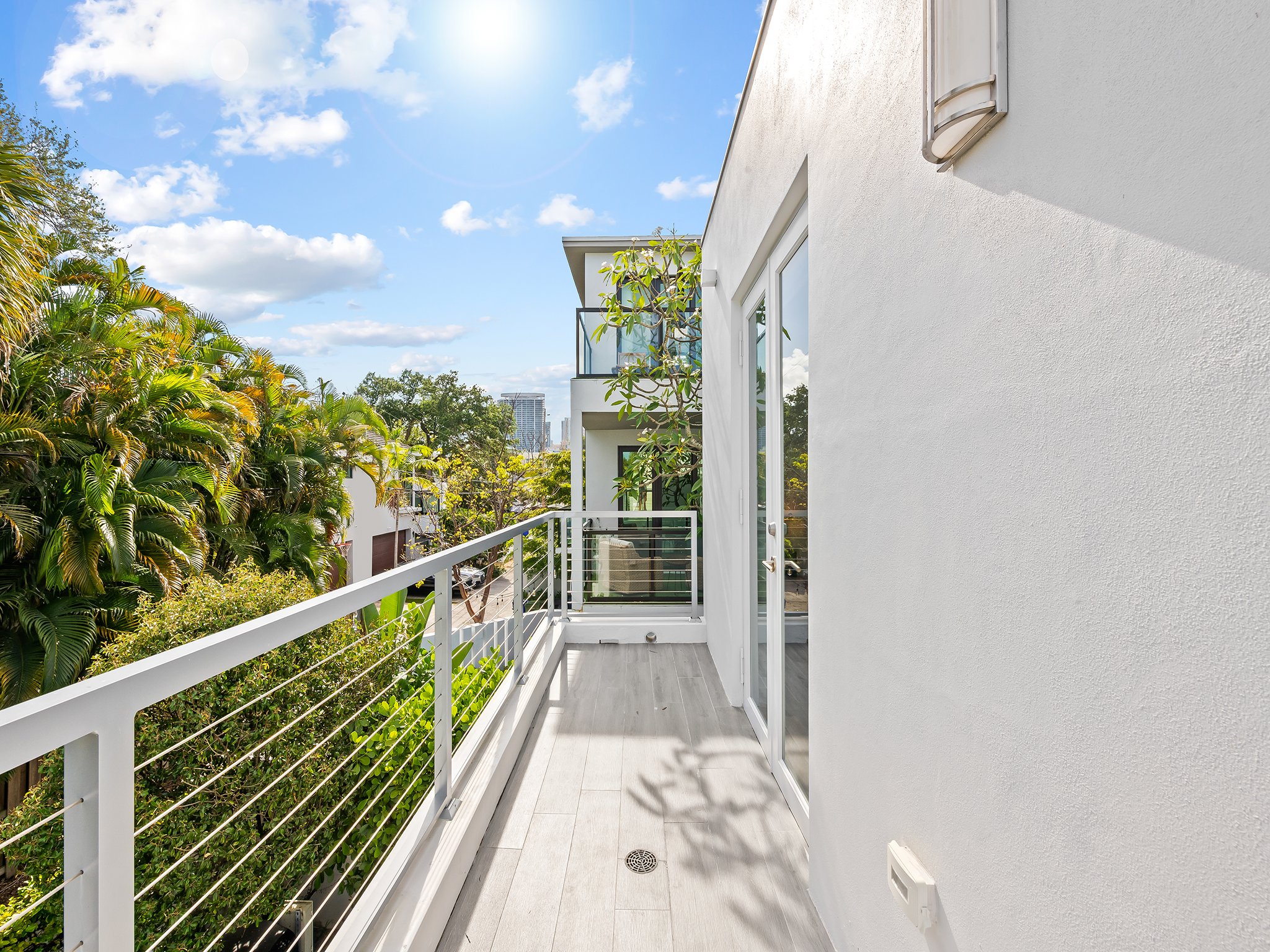 A balcony with a white railing and a sliding glass door, overlooking trees and a cityscape under a partly cloudy sky.