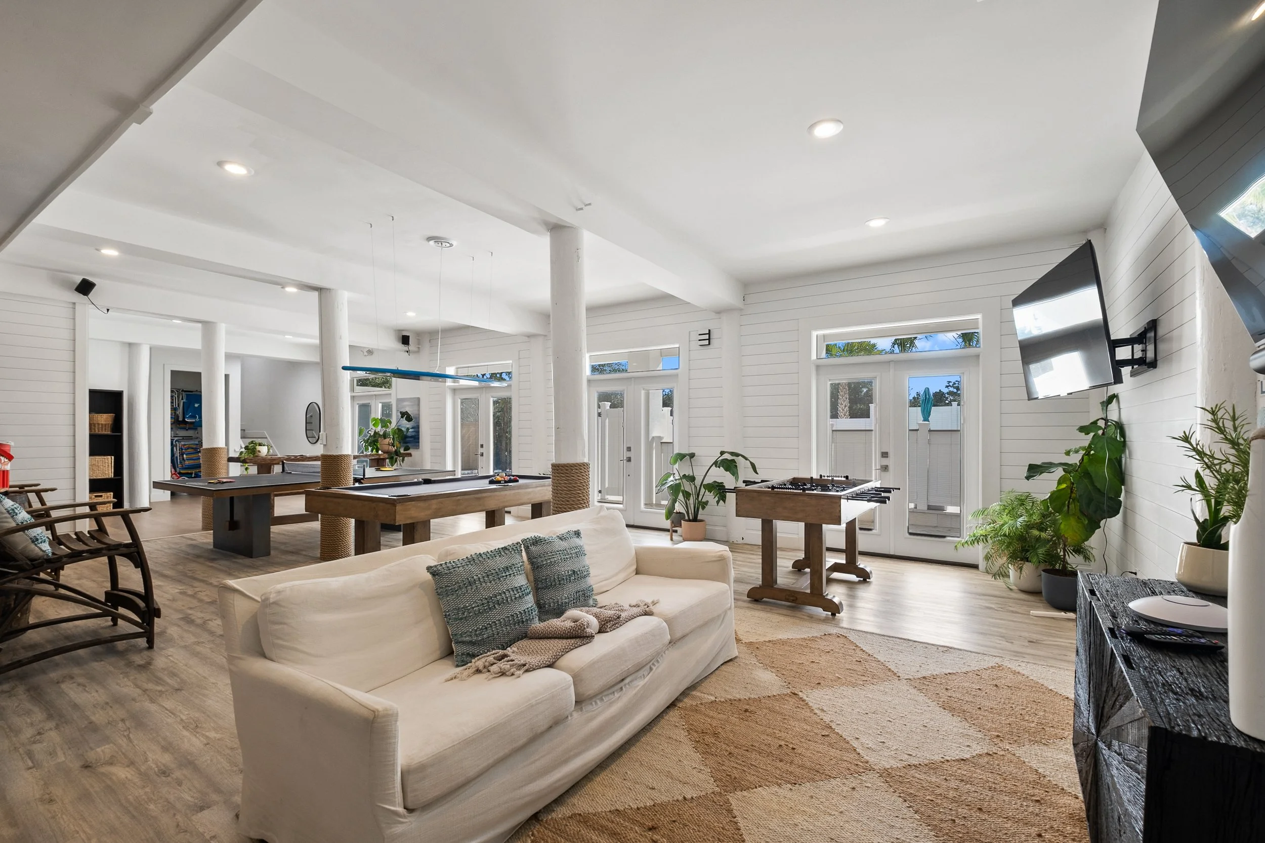 Spacious living room with white walls and wooden floors, featuring a white sofa with blue pillows, several potted plants, large windows and glass doors, a wooden gaming table, and mounted flat-screen televisions.