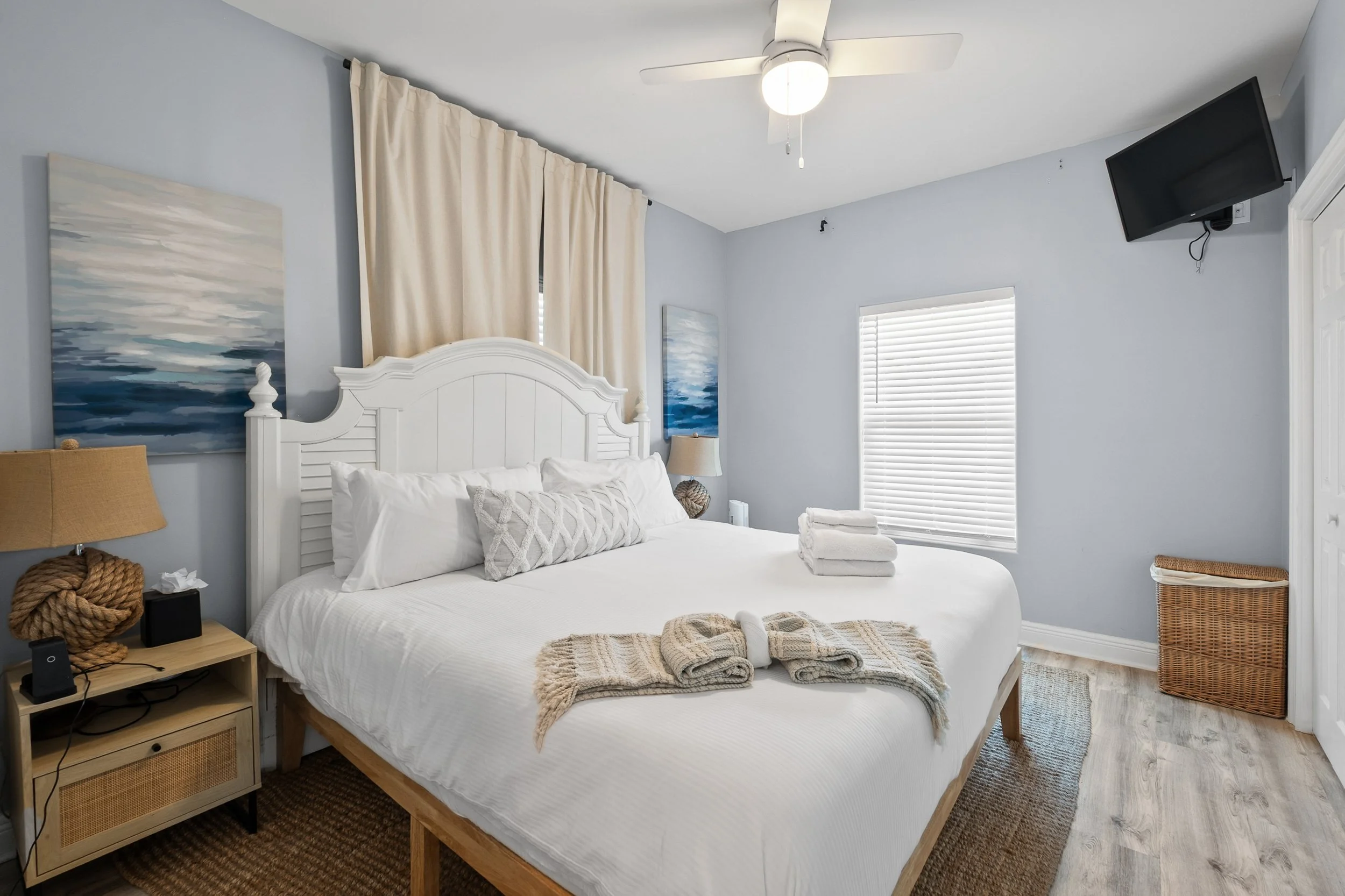 Clean and cozy bedroom with a white bed, beige curtains, paintings of ocean scenes, a ceiling fan, and a small TV mounted on the wall.