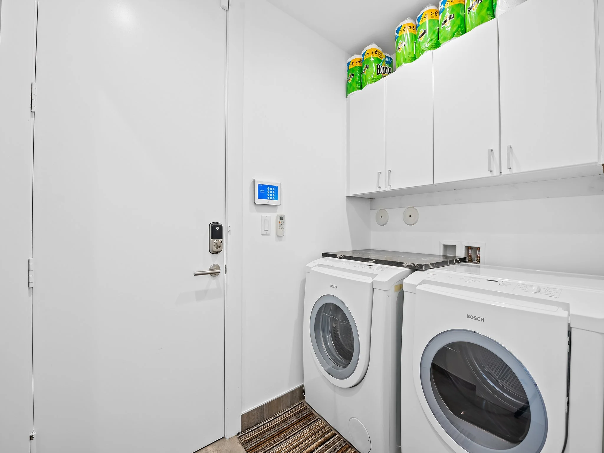 A laundry room with white cabinets and walls, two front-loading Bosch washers, a countertop, and a shelf with paper towels. There is a digital control panel on the wall and a freshener dispenser.