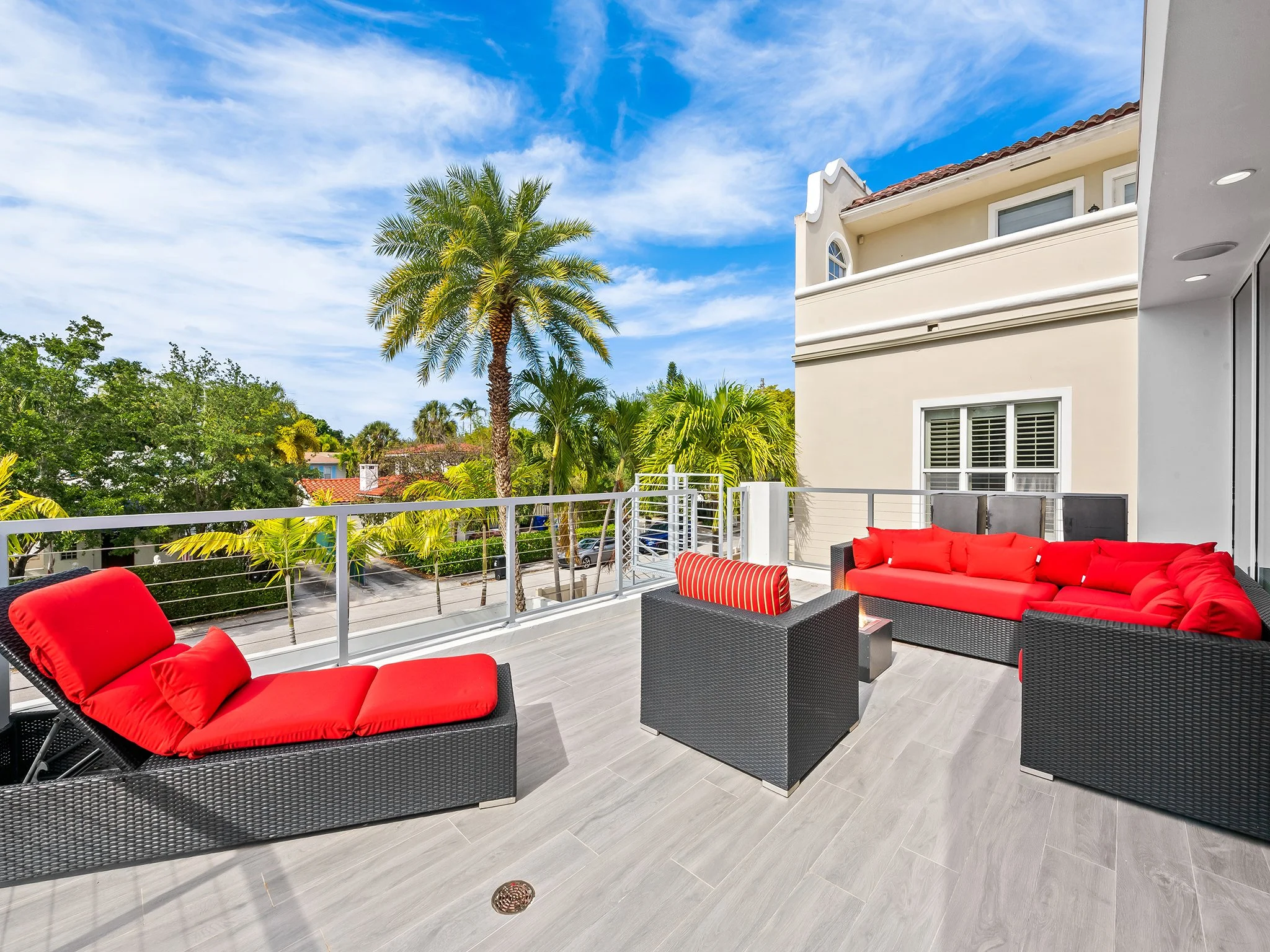 A spacious balcony with a modern outdoor seating area featuring black rattan furniture with red cushions. The balcony has gray wood-look tile flooring and a white railing, overlooking lush green trees, including a prominent tall palm tree. The backgr