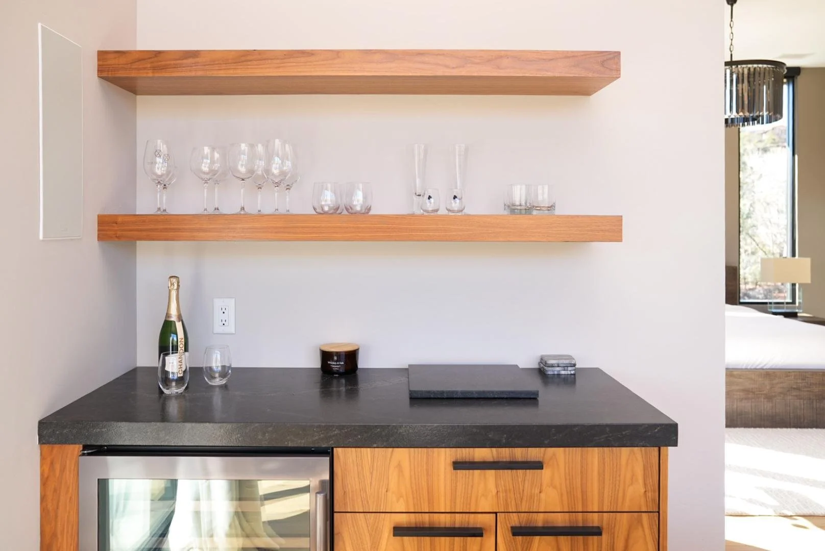 A kitchen area with a black stone countertop, wooden cabinets, and open wooden shelves holding wine glasses, champagne flutes, and other glassware. Part of a wine fridge is visible, along with a black candle jar and a small box of coasters.