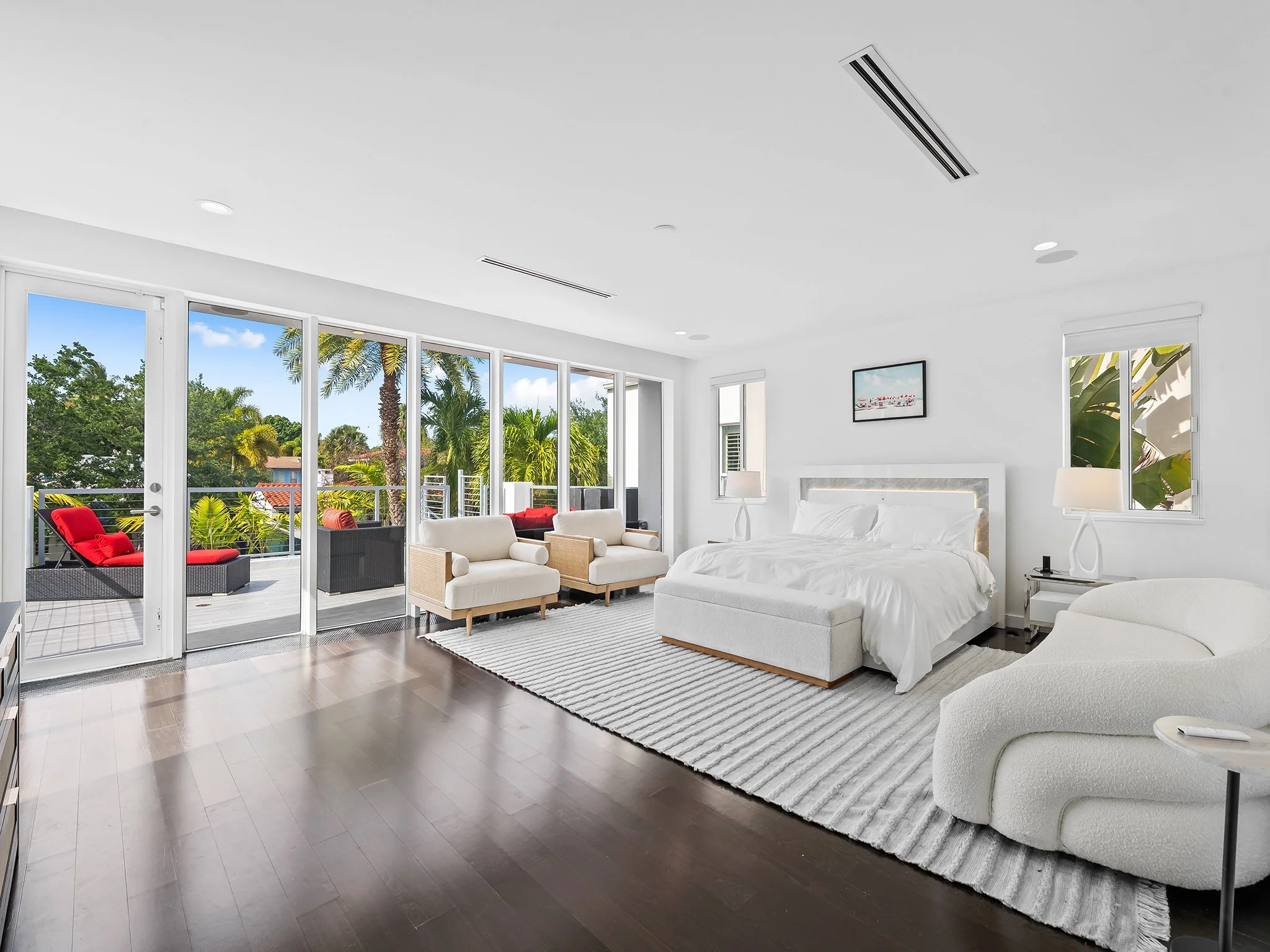 Bright living room with large windows and sliding glass door opening to balcony with outdoor seating, palm trees visible outside, white walls, modern furniture, ceiling with air vents, framed artwork, and area rug.