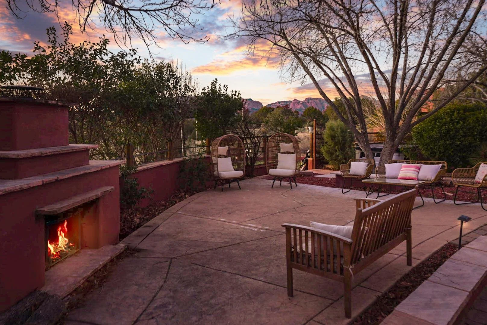A cozy outdoor patio at sunset with a small fireplace, various chairs, a wooden bench, and a view of distant mountains and trees.