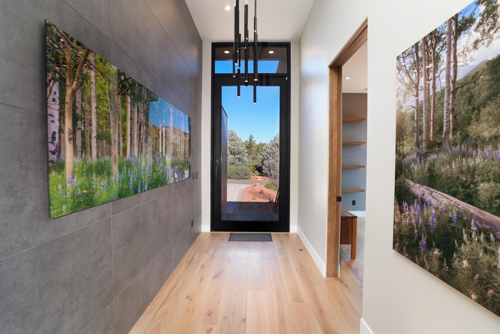 Modern entryway with wooden floors, large landscape photographs on the walls, and a glass door leading outside to a garden and clear blue sky.