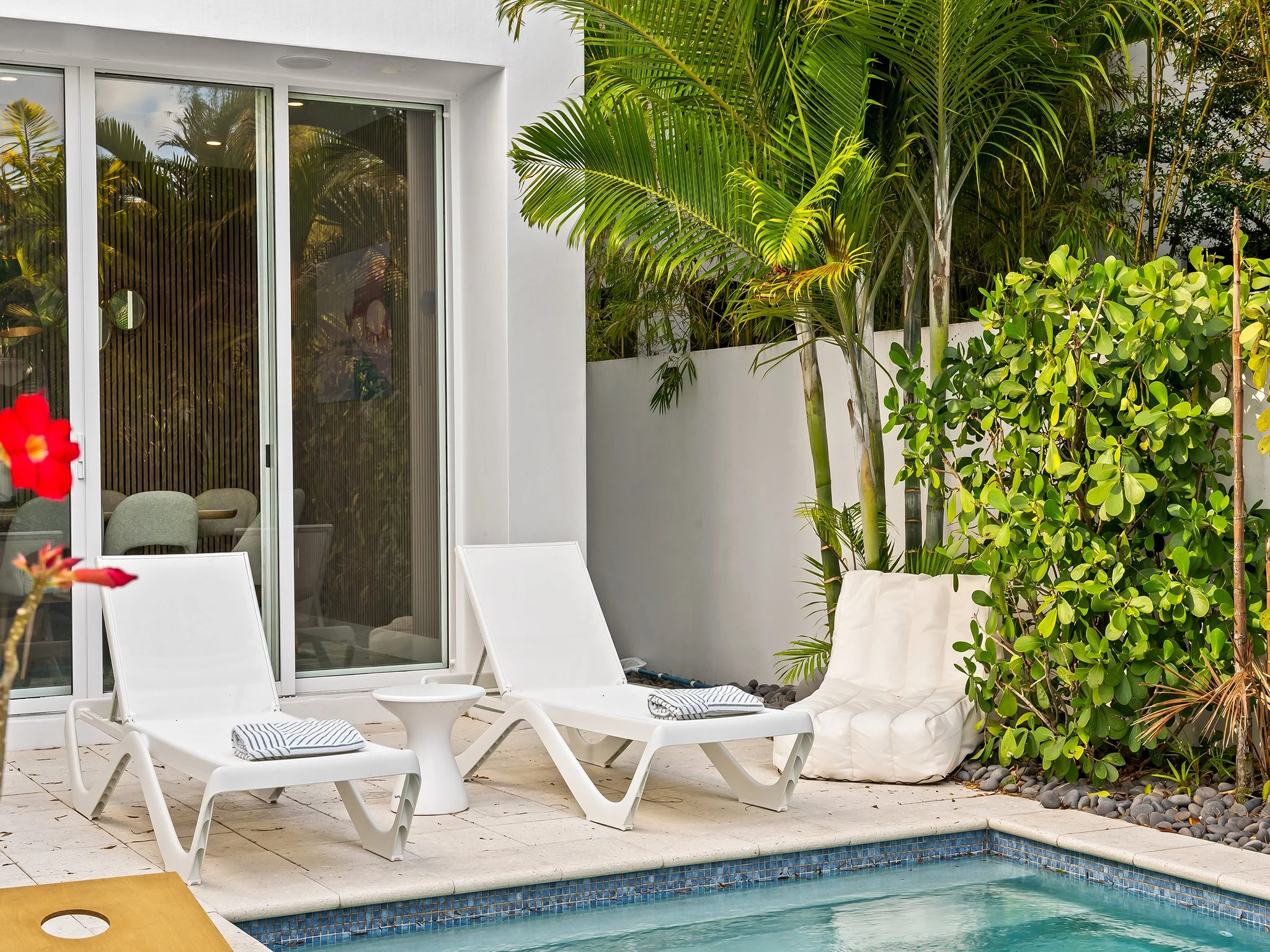 Poolside area with white lounge chairs, a small white table, greenery including palm trees and shrubs, a white couch, and a glass sliding door leading indoors.