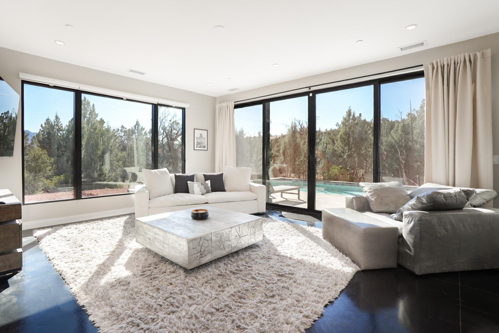 Bright living room with large windows, white sofas, a shaggy rug, and a view of a backyard with a pool and trees