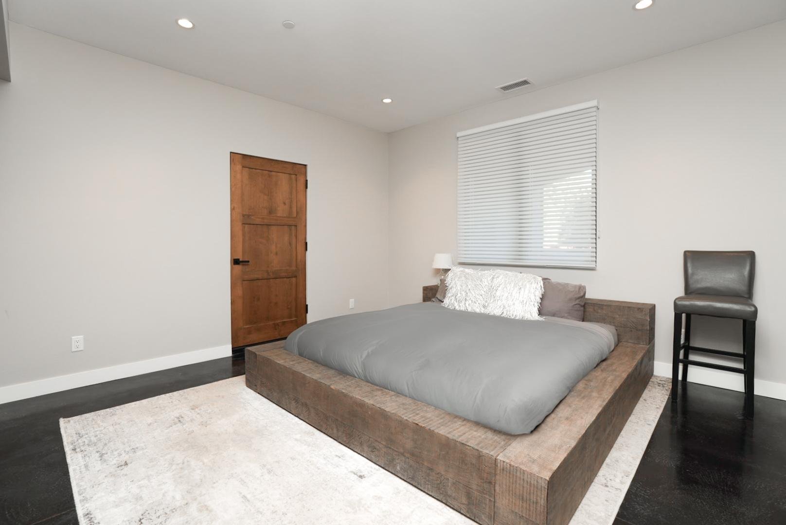 Minimalist bedroom with a large wooden bed frame, grey bedding, white pillows, a side lamp, a window with white blinds, a wooden door, and a tall grey chair.