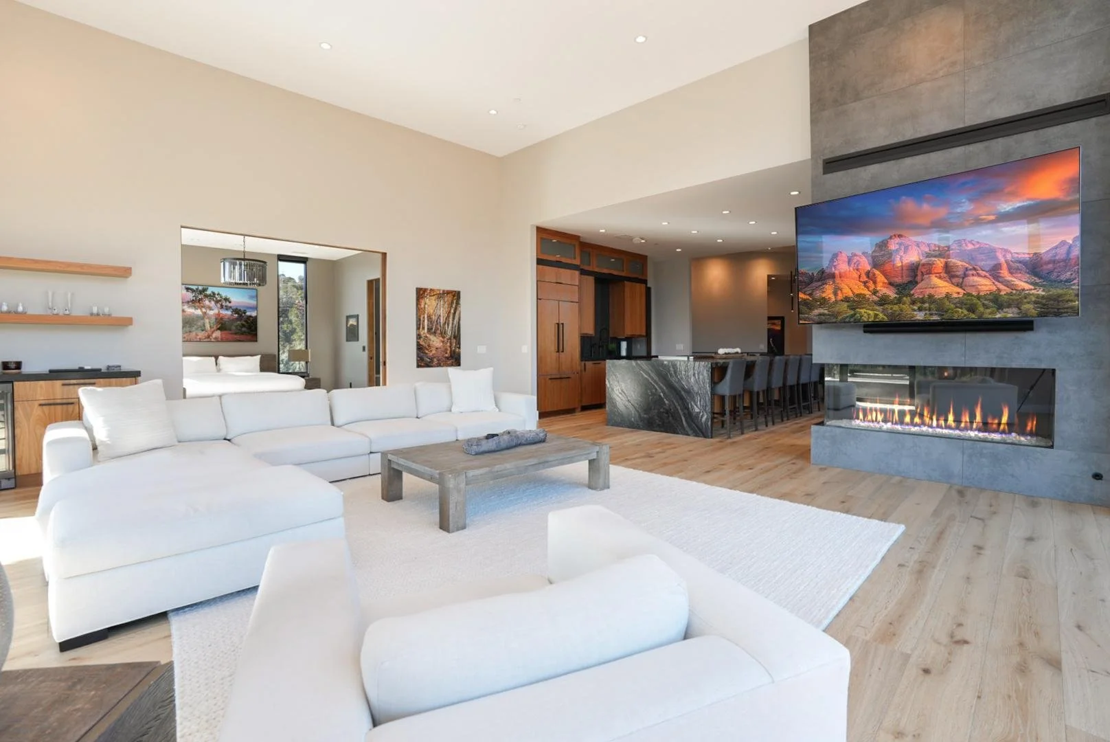 Modern open-concept living room with white sectional sofa, wooden coffee table, large wall-mounted flat-screen TV, fireplace, and a bar area with stools, with a bedroom visible through an open doorway.