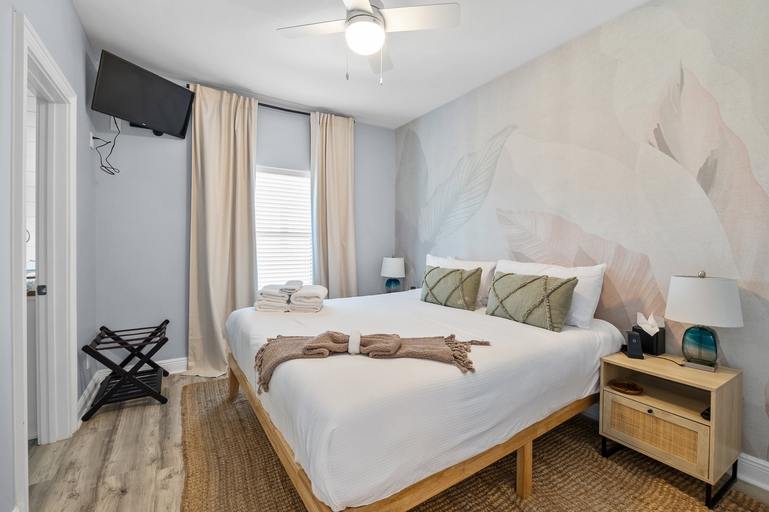 A bedroom with a large bed, pillows, and blankets, a nightstand with a lamp and tissue box, a wall with a floral mural, a window with curtains, a ceiling fan, and a small TV mounted on the wall.