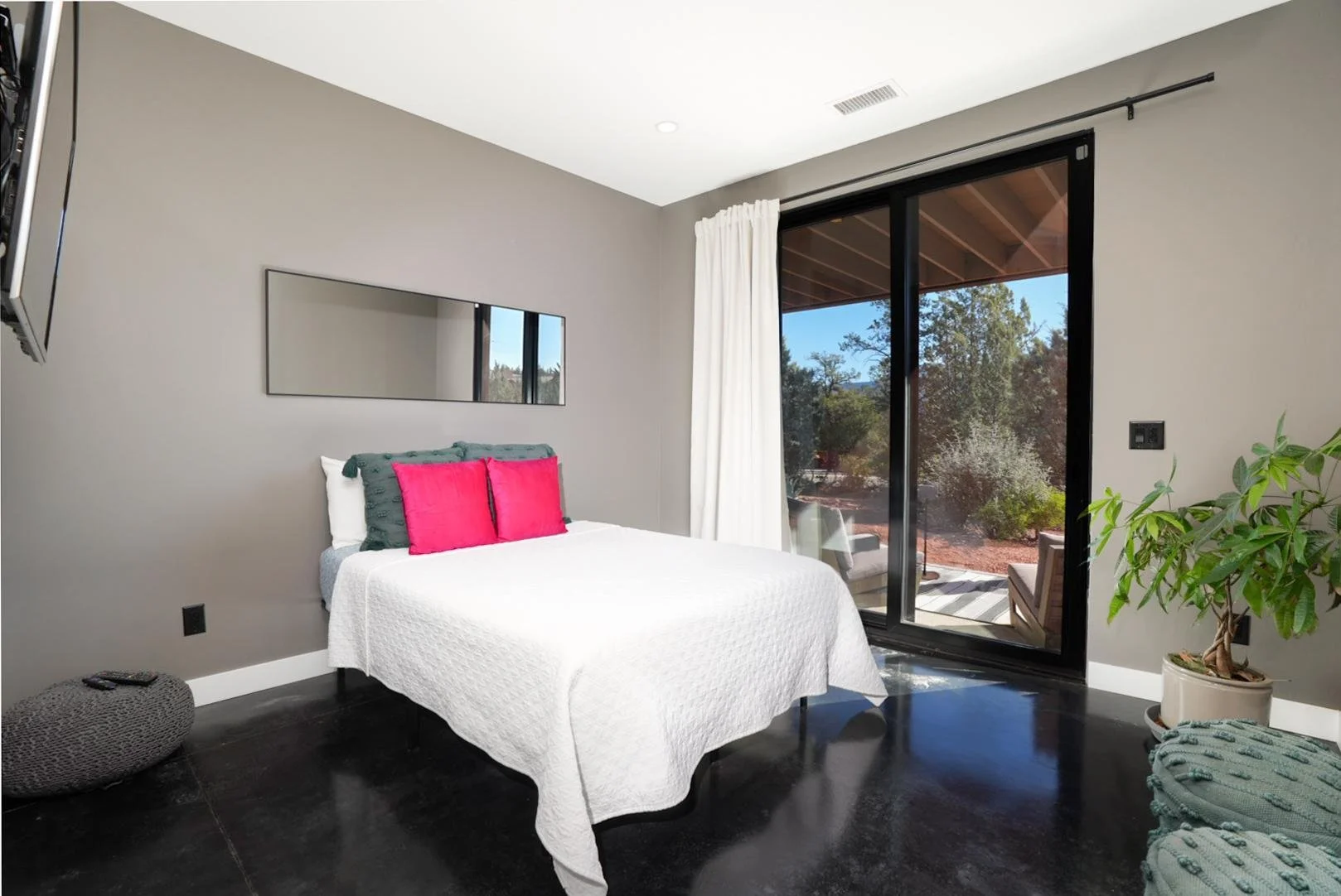 A bedroom with a bed covered in a white quilt, two pink pillows, and dark green pillows, a large sliding glass door with white curtains, and a window reflected in a wall mirror, with outdoor patio furniture visible through the door.