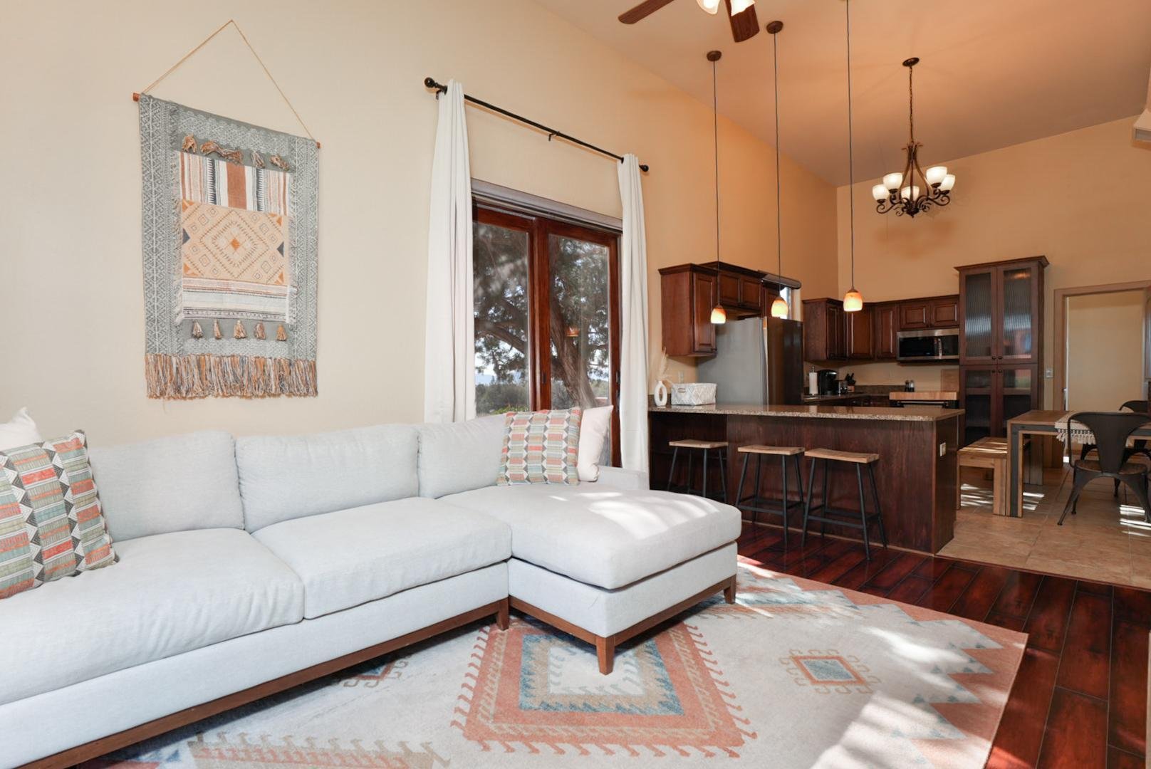 Living room with white sectional sofa, patterned pillows, large window with white curtains, wall hanging, and open kitchen with wooden cabinets, island with stools, and dining table in background.