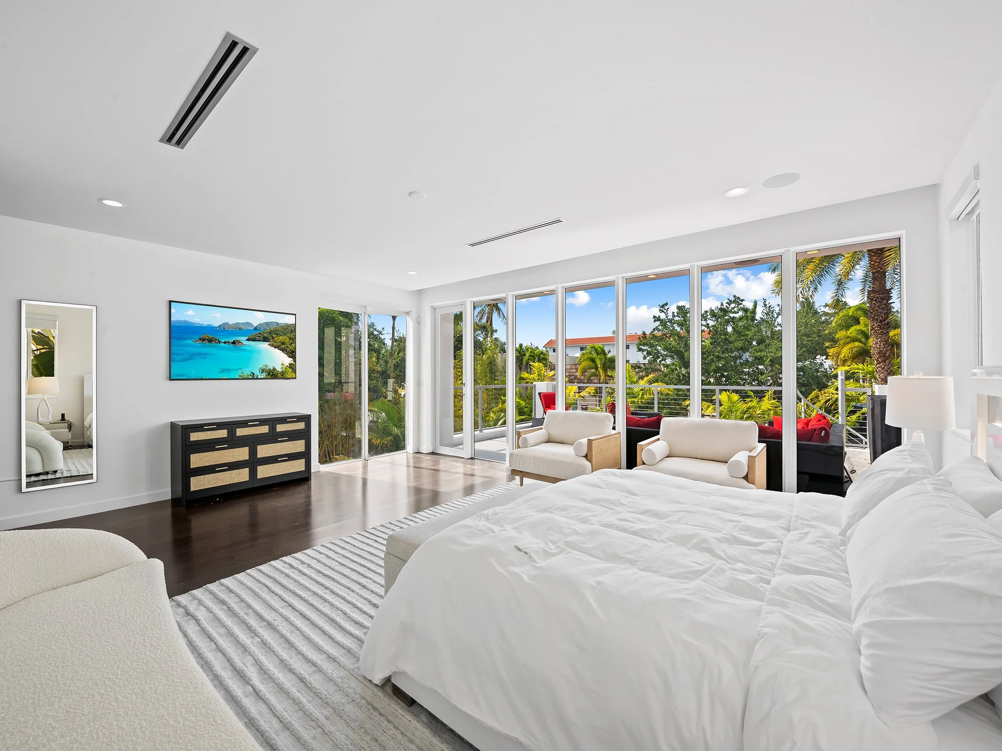 Modern living room with large windows showing a tropical landscape, white furniture, and a flat-screen TV on the wall.