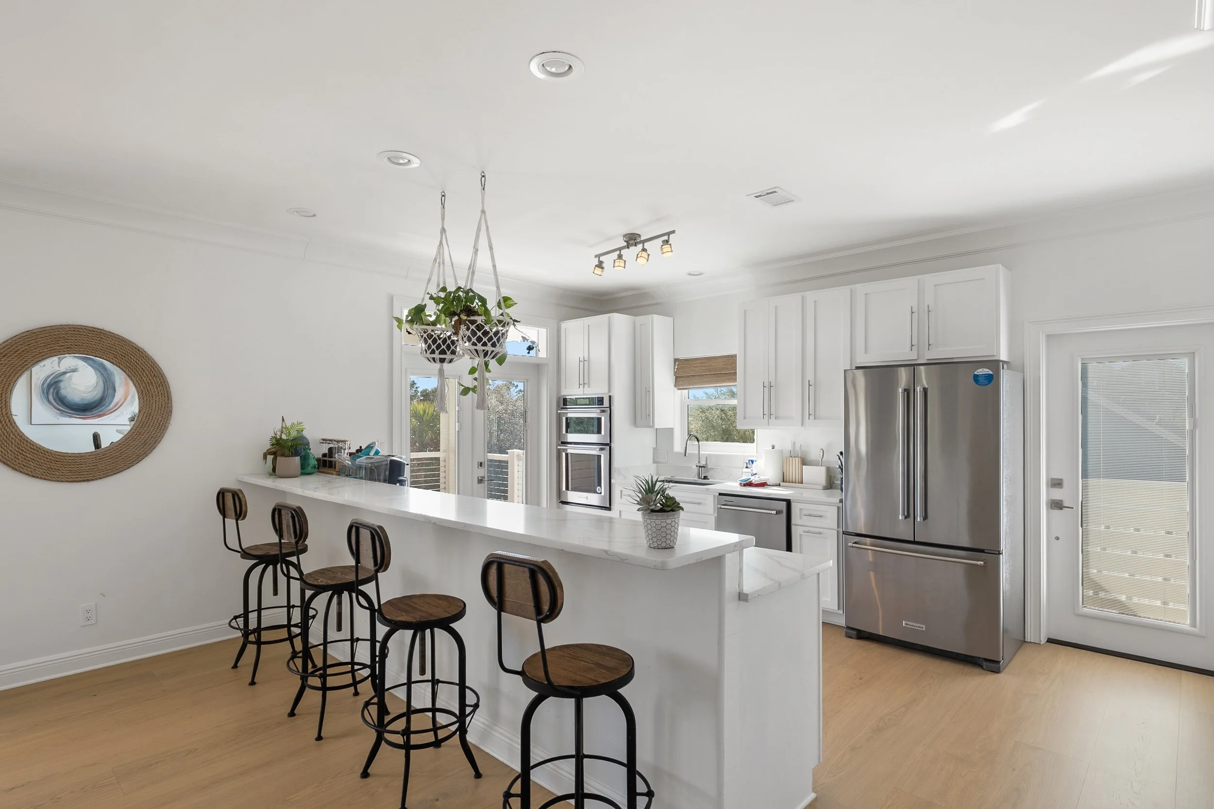 Bright modern kitchen with white cabinetry, stainless steel appliances, a breakfast bar with four wooden stools, hanging plants above the bar, and hardwood floors.