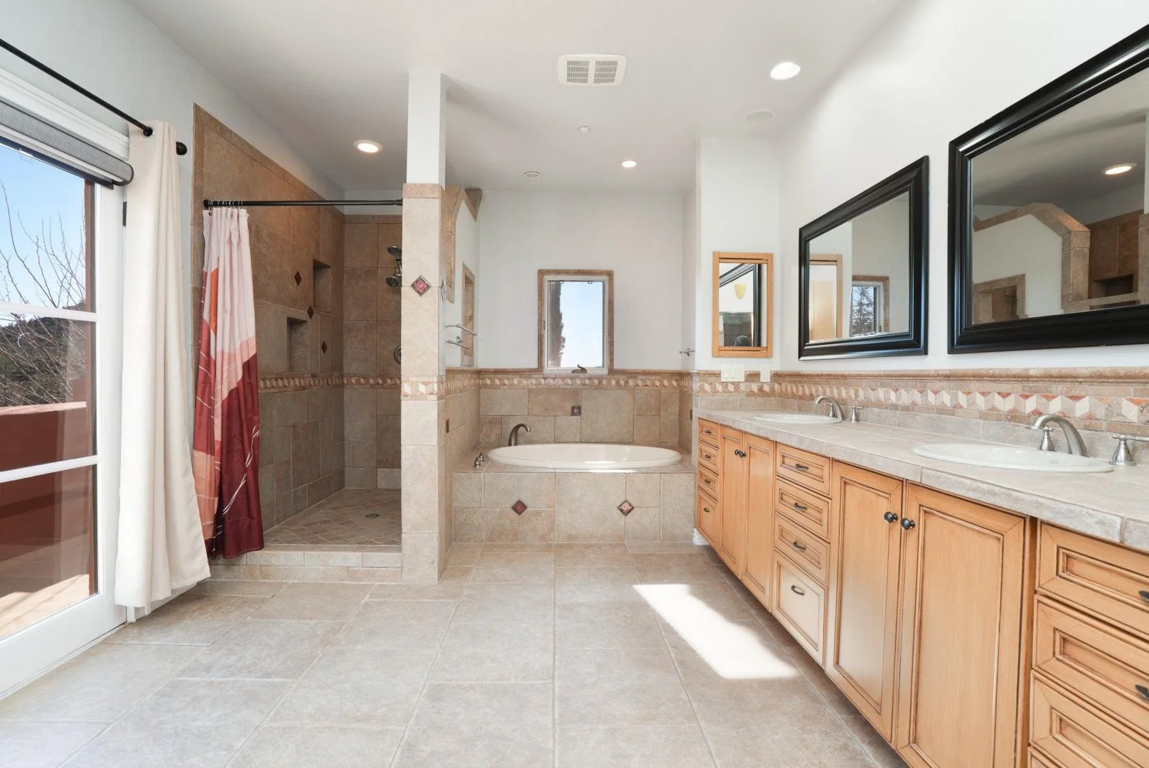 A spacious bathroom with a glass door leading outside, a walk-in shower, a bathtub beneath a small window, a double sink vanity with wooden cabinets, large mirrors, and beige tile flooring and walls.