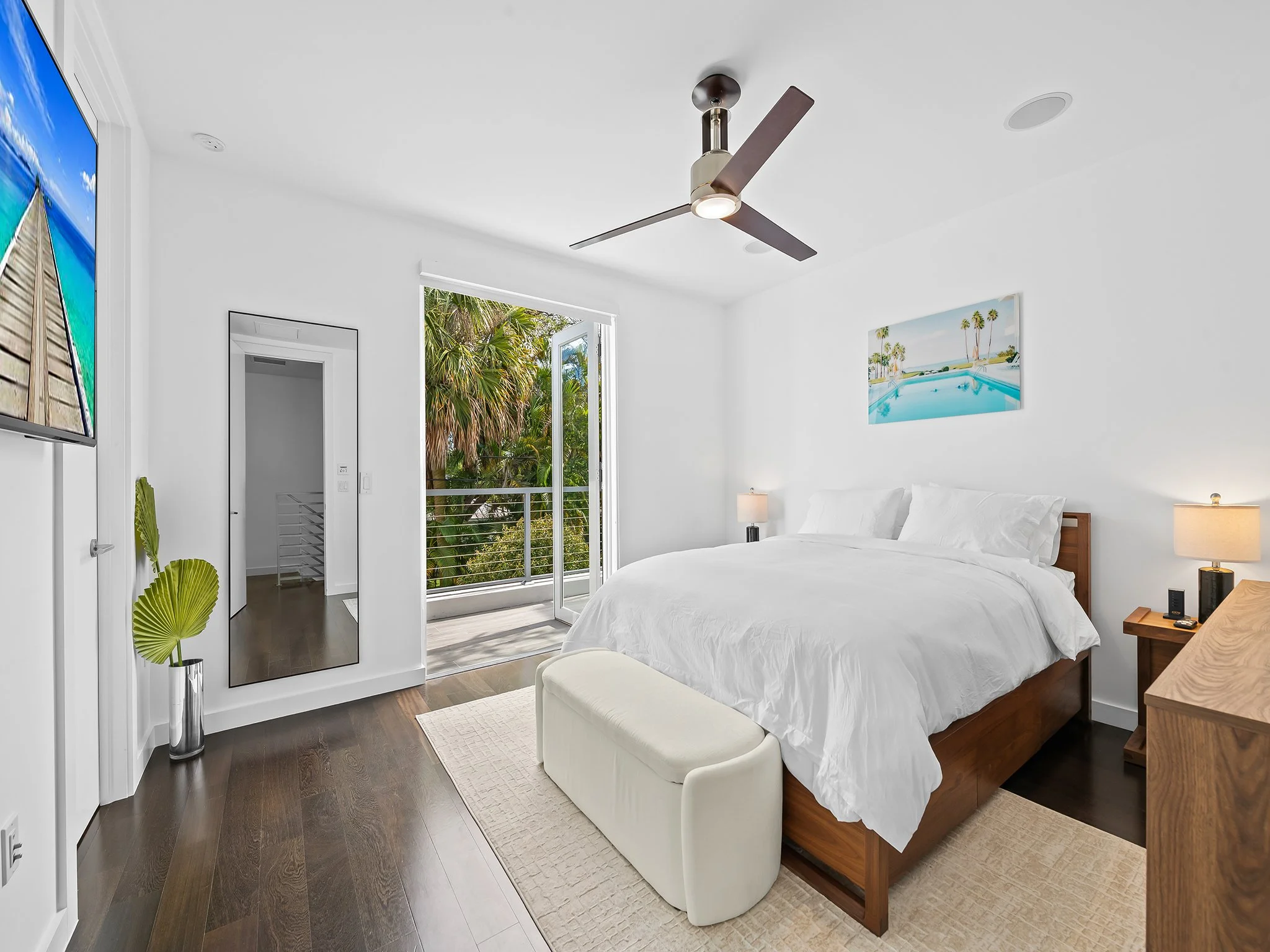 A modern bedroom with white walls, a ceiling fan, a large window with a sliding door, a wall-mounted TV, a picture of a pool scene, a bed with white bedding, a bench at the foot of the bed, a wooden side table, a lamp, and a full-length mirror.