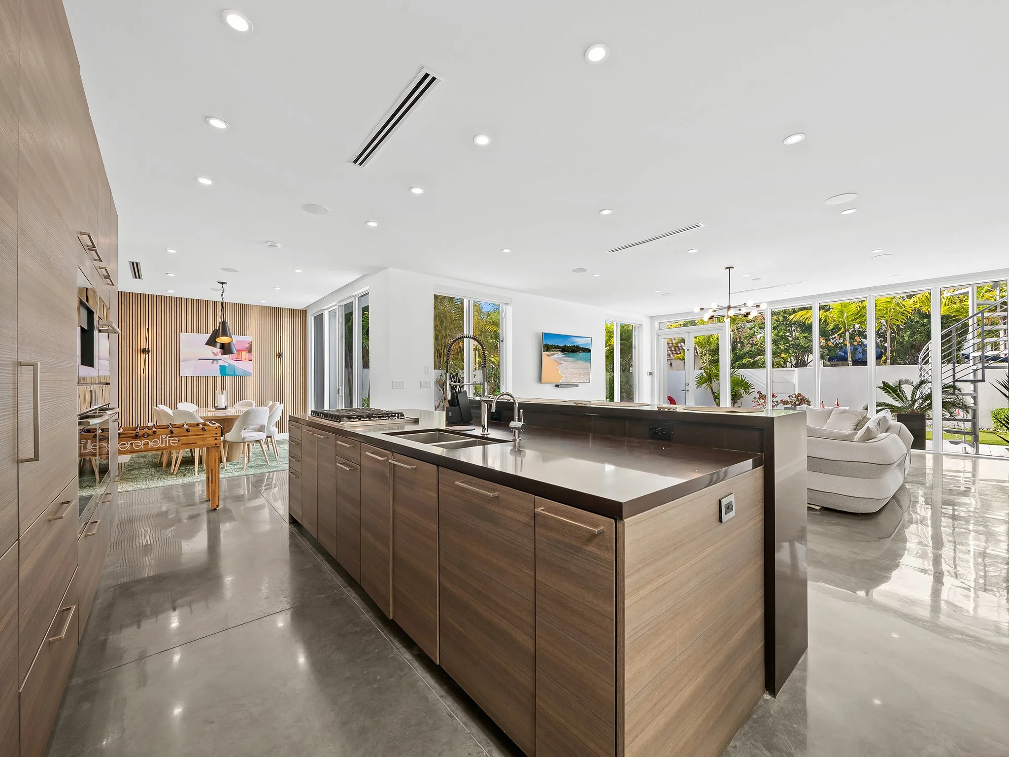 Modern open-plan kitchen with wooden island, bright natural light, large windows showing a backyard garden, contemporary furniture, and a living area.