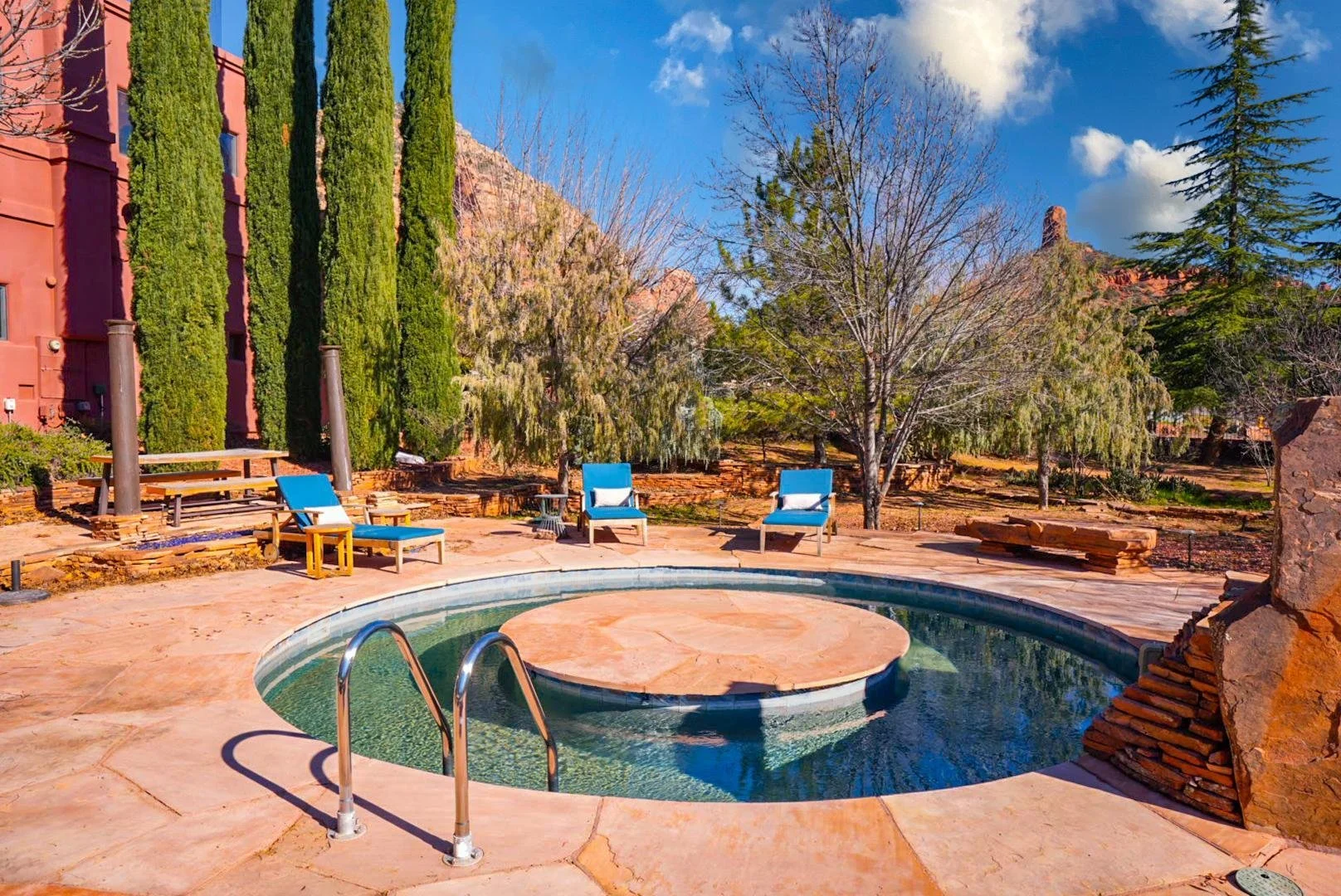 Outdoor scene with a small circular pool, blue lounge chairs, benches, and tall trees under a blue sky with clouds.
