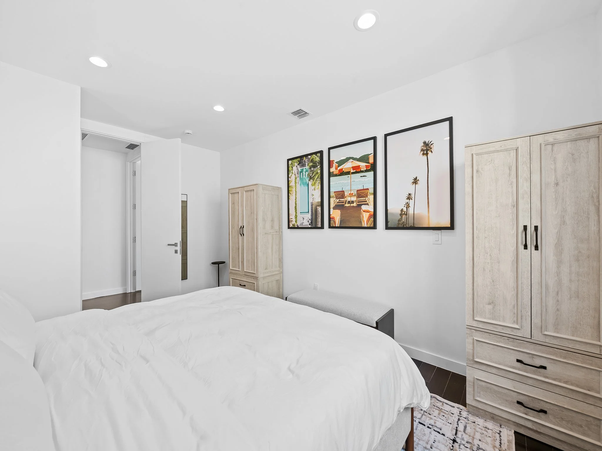 A minimalistic bedroom with white walls and ceiling, featuring modern recessed lighting, framed beach-themed artwork, light wood cabinets, and a white bed with a tray ceiling.