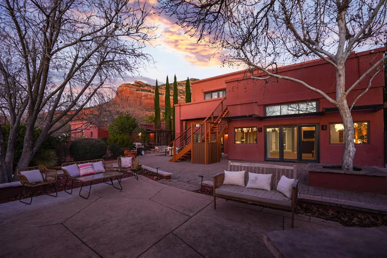A backyard patio at sunset with outdoor furniture, trees, and a reddish house with stairs, against a mountainous landscape with a pink and purple sky.