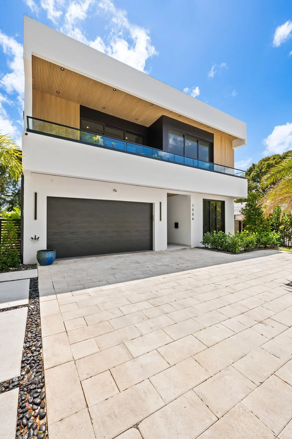 Modern white two-story house with black accents, a gray garage door, a balcony with glass railing, and lush green landscaping against a bright blue sky.