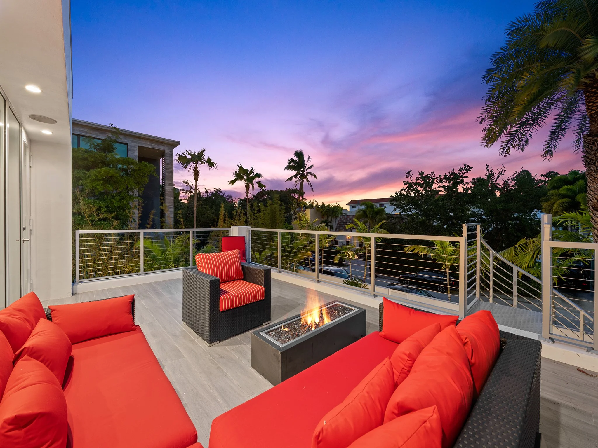 A modern outdoor balcony with red cushioned seating, a fire pit, and palm trees during sunset