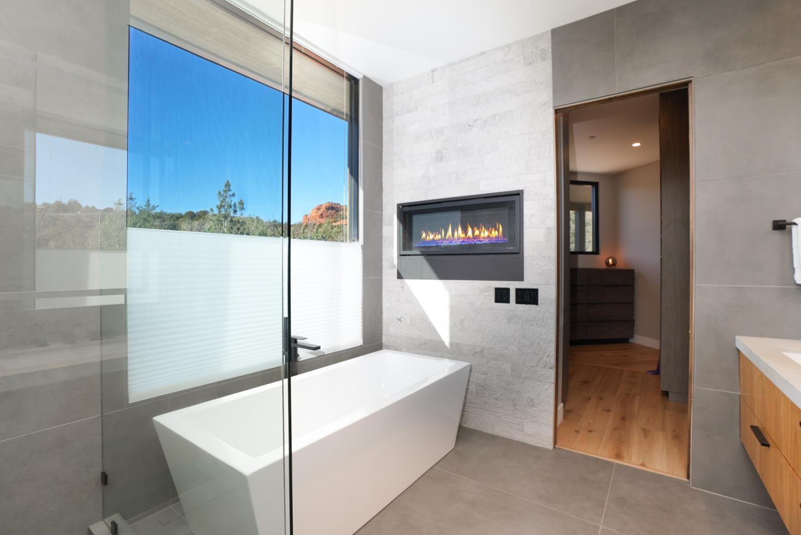 Modern bathroom with a freestanding bathtub, large window with a view of trees and a red rock, wall-mounted gas fireplace, and wooden flooring visible through an open door.