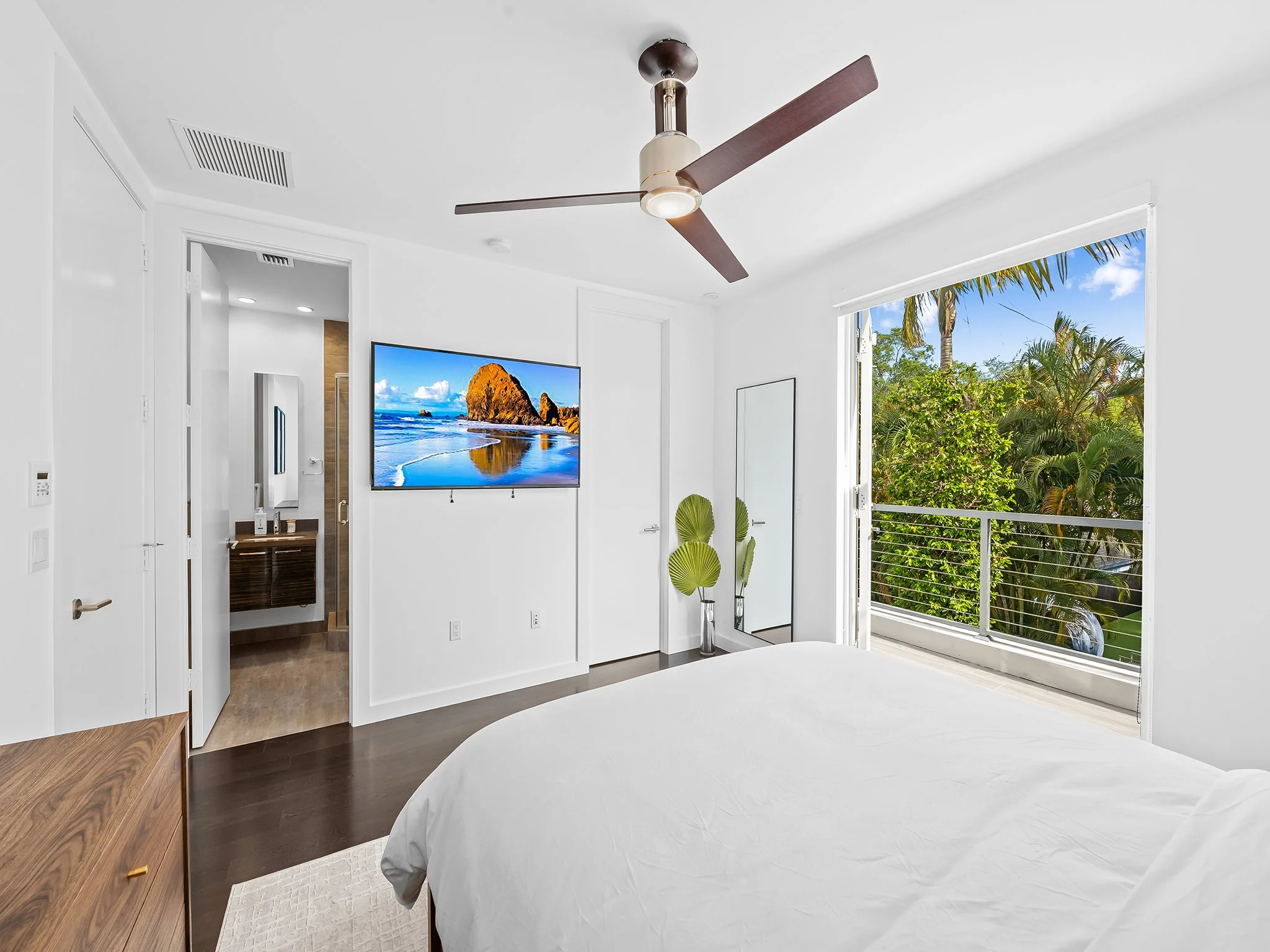 A bright bedroom with a white bedspread, a ceiling fan, and large open window showing trees and blue sky.