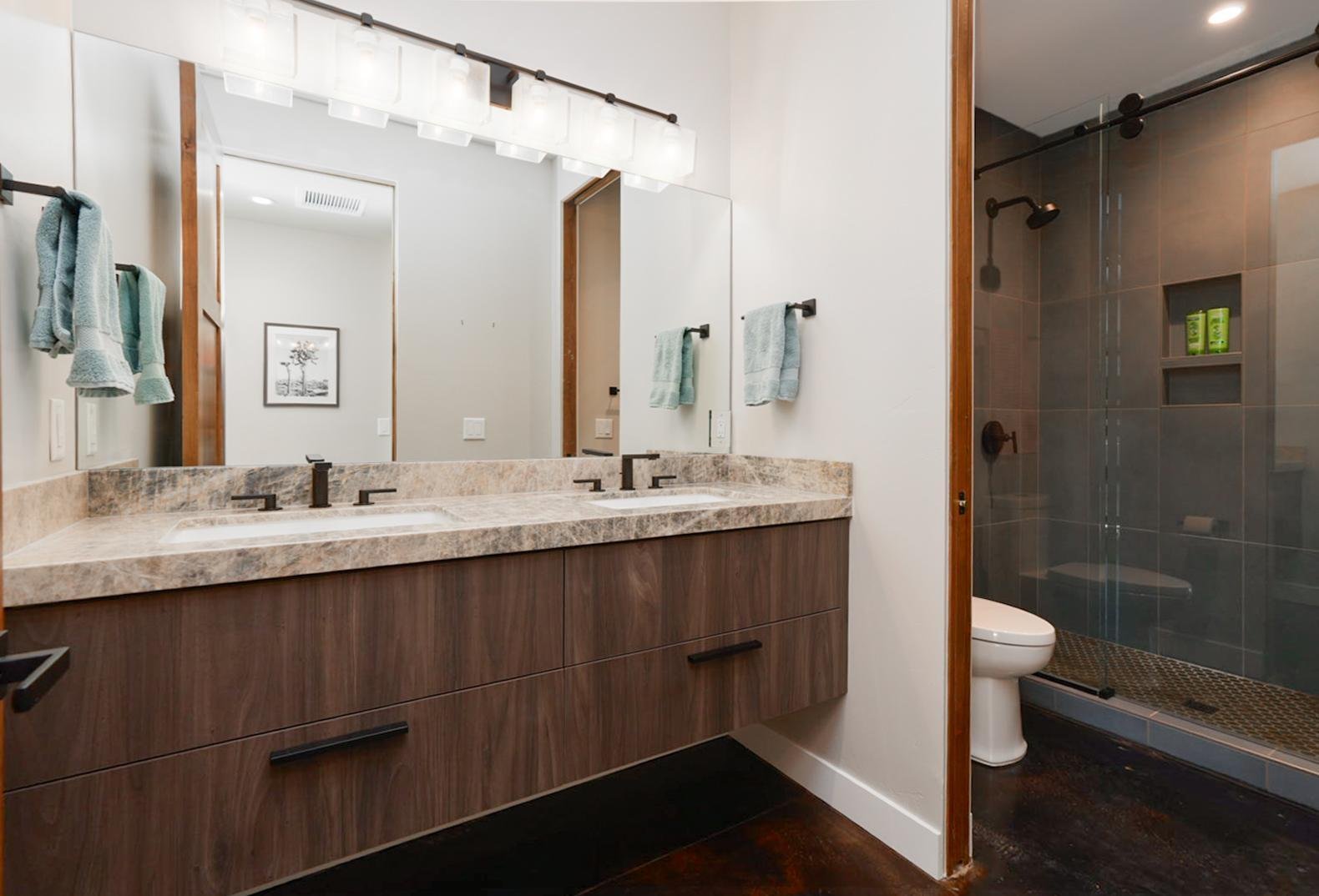 Modern bathroom with double vanity with marble countertop and wood cabinet, large mirror, towel bars with teal towels, shower with glass door, tile walls, and built-in shower niche.