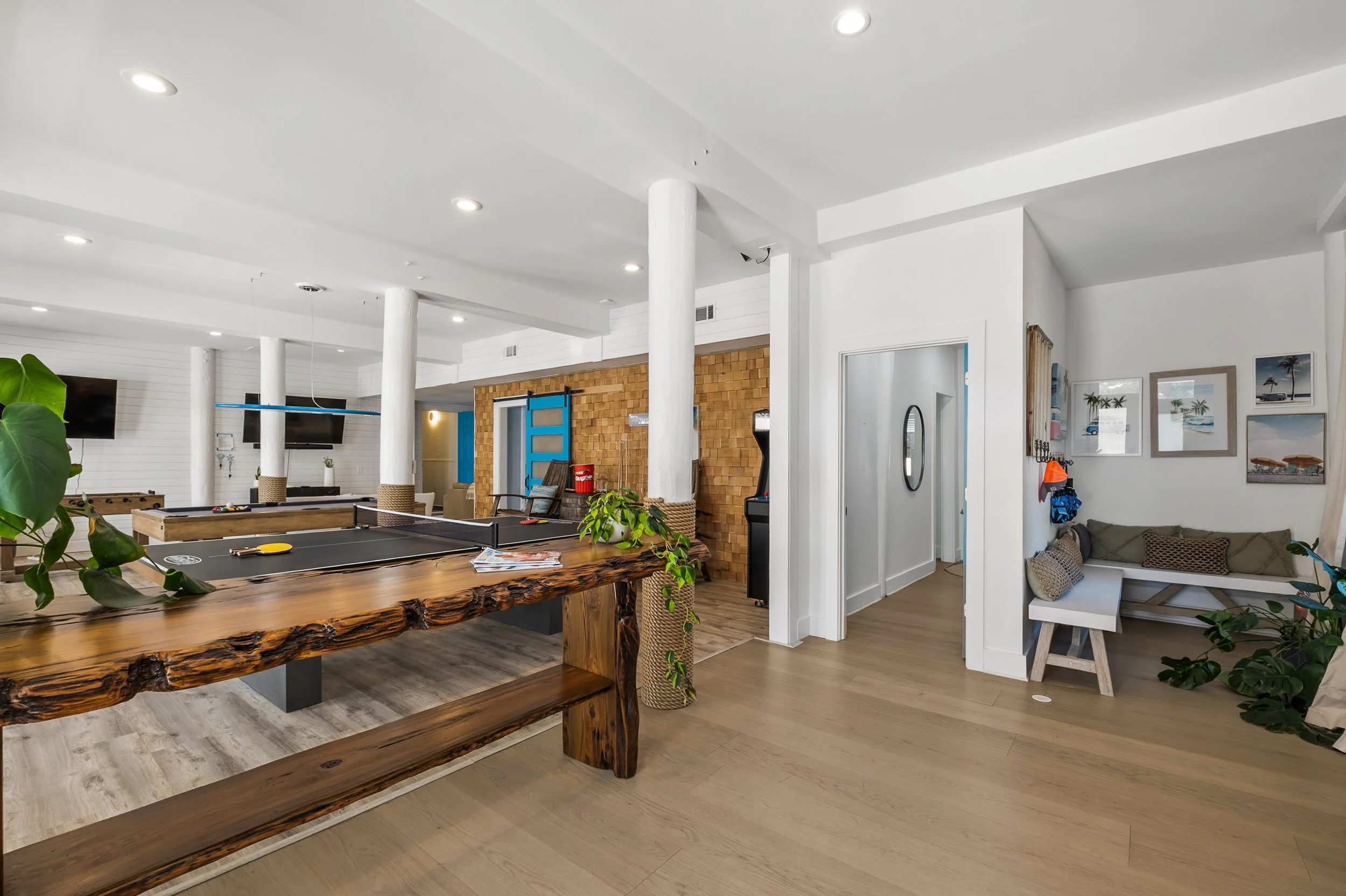 Interior view of a modern recreational space with a wooden reception desk, a ping pong table, multiple wall-mounted TVs, and beach-themed artwork. There are white support columns, wood flooring, and a small sitting area with cushions.