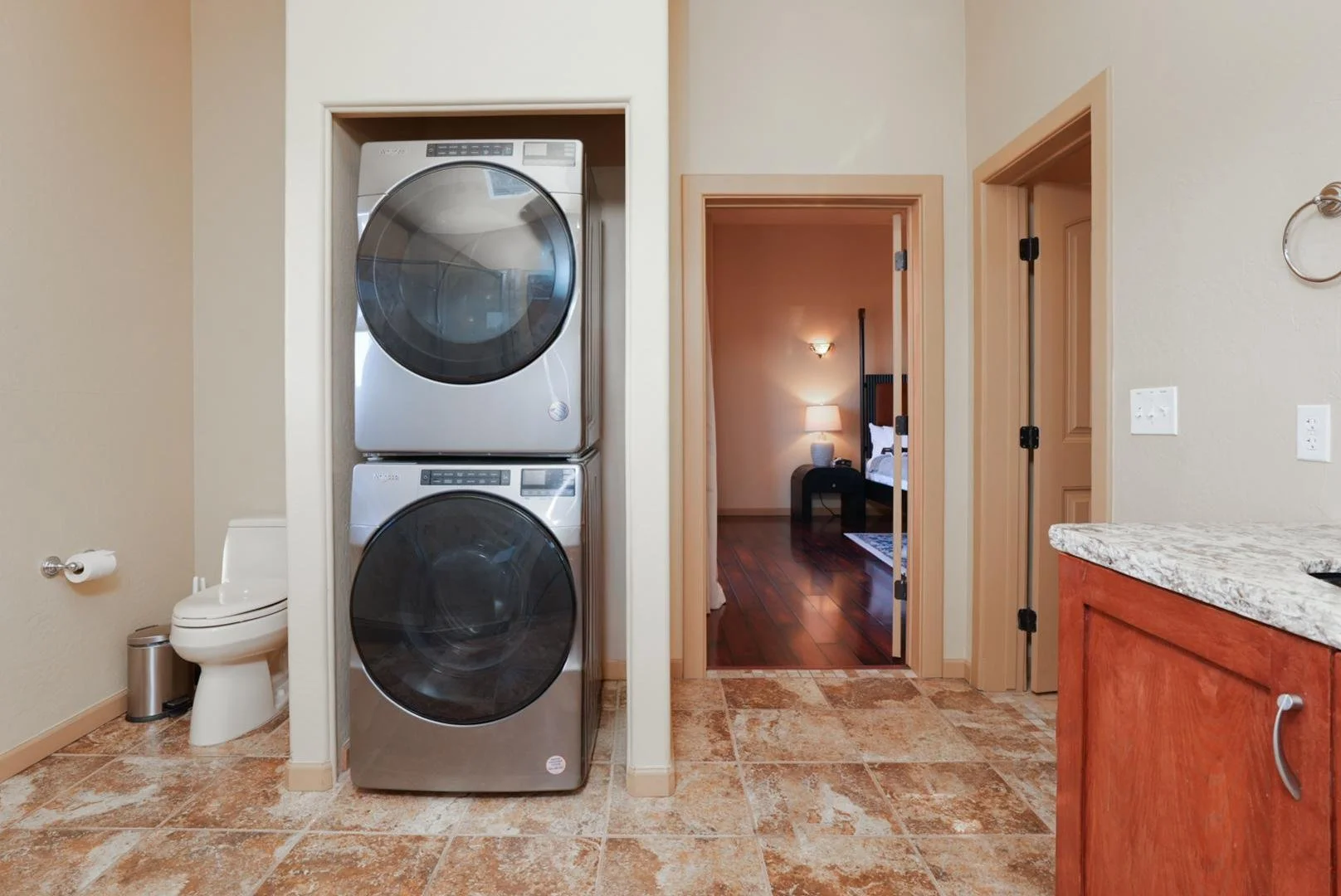 A laundry area with a stacked washer and dryer, a toilet, and a toilet paper holder. There is a doorway leading to a bedroom with a nightstand, lamp, and bed, and another doorway with a door frame. The floor is tiled, and a wooden cabinet with a gran