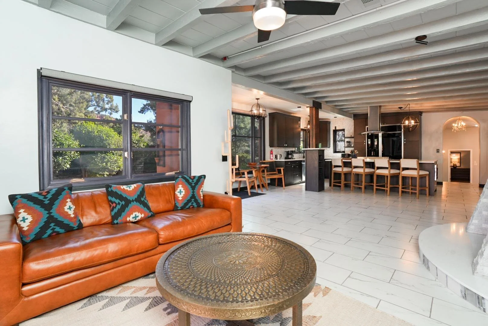 Open-concept living room and kitchen with a brown leather sofa, colorful cushions, large windows, tiled floor, black kitchen cabinets, a breakfast bar with chairs, and modern lighting fixtures.