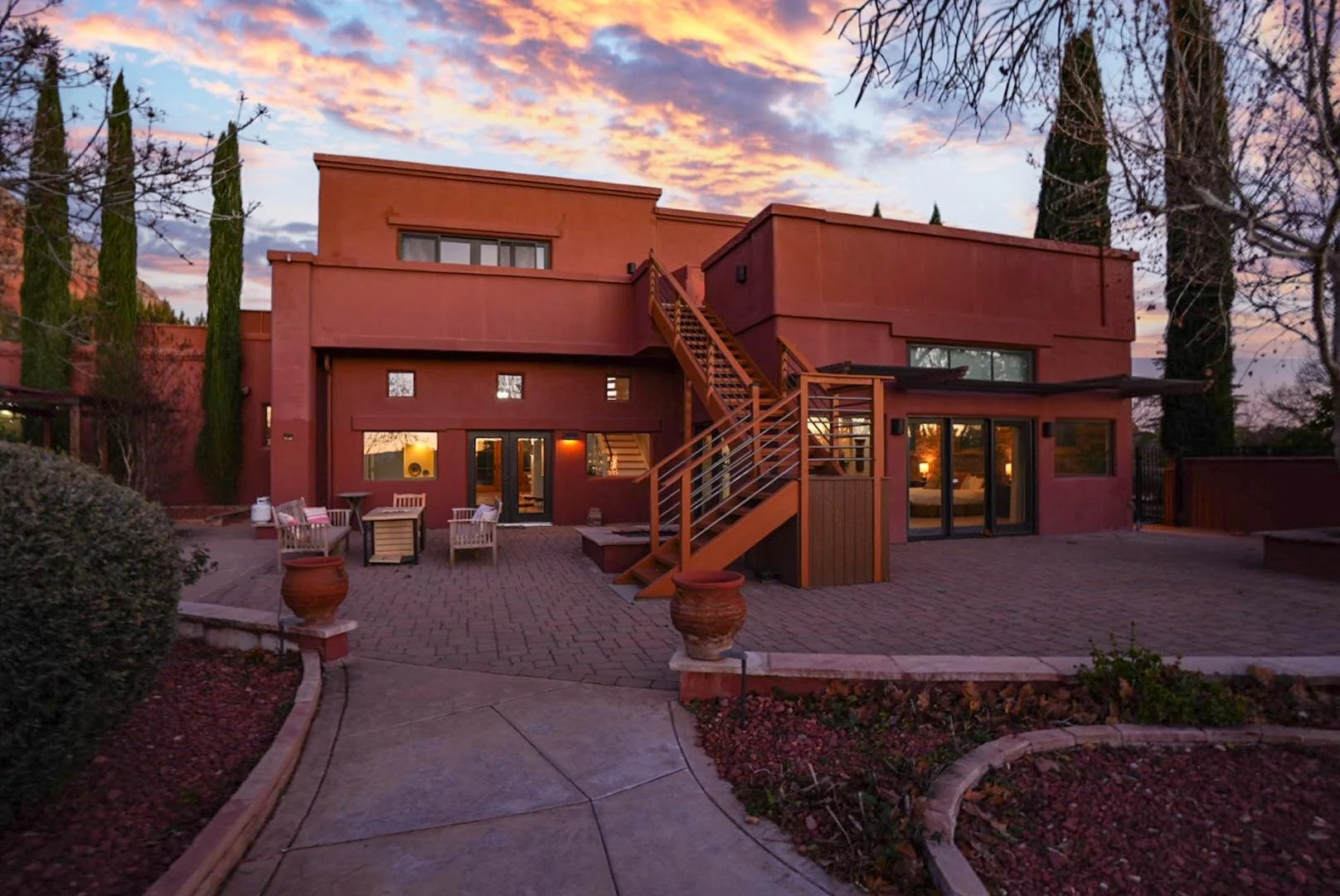 The back of a two-story house with a patio at sunset, featuring a staircase, outdoor seating, potted plants, and large glass sliding doors.