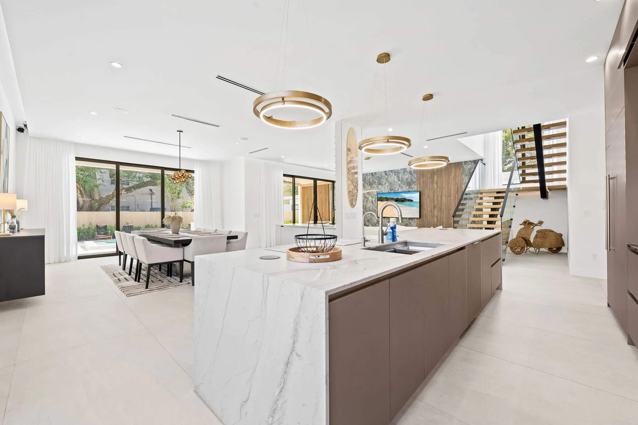 Open concept modern kitchen and dining area with large island, marble countertops, pendant and circular ceiling lights, wood and marble accent walls, sliding glass doors overlooking the backyard, and staircase with wooden steps and metal railing.