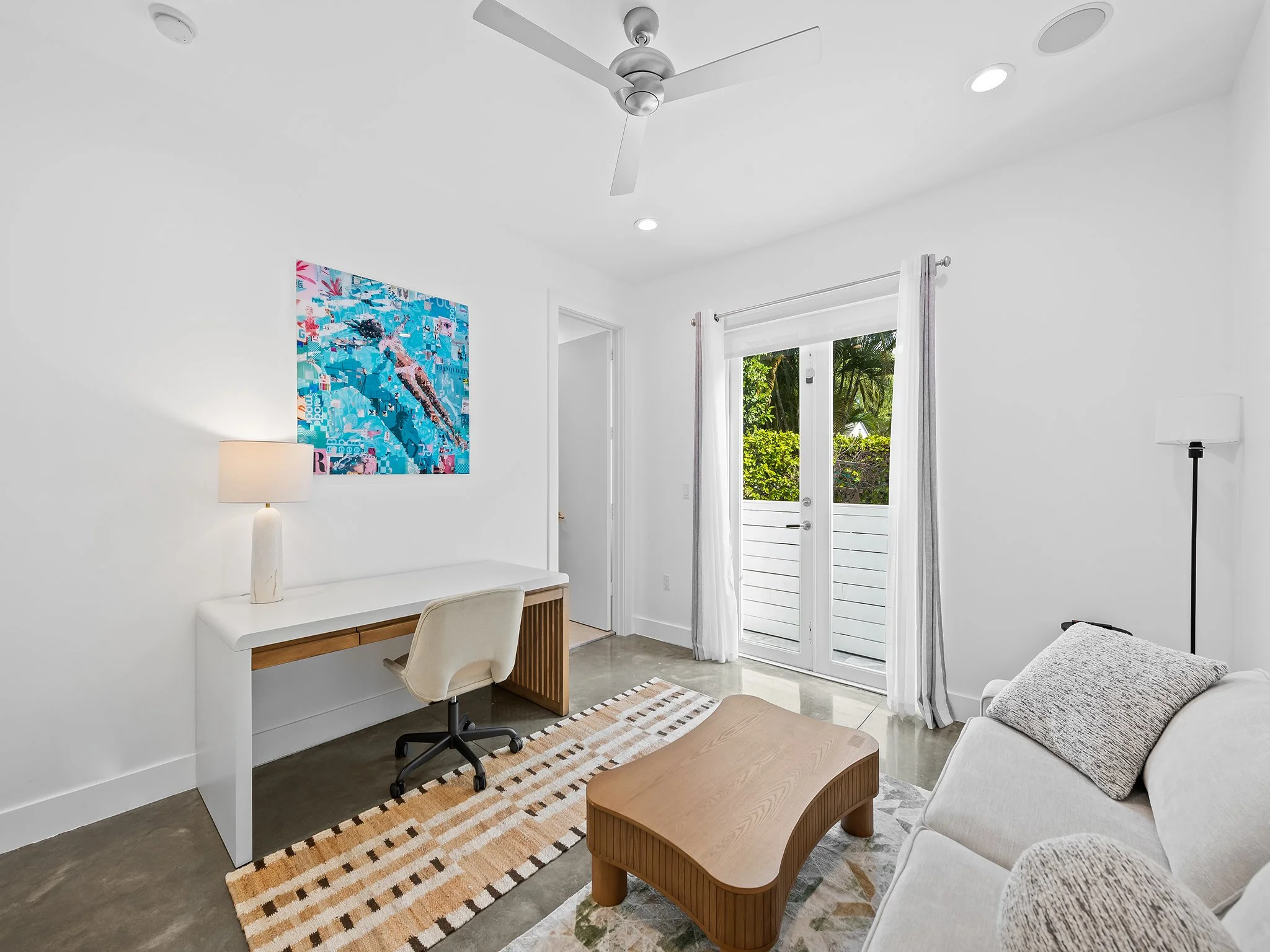 A bright, white room with a ceiling fan, recessed lighting, and a double door leading outside. The room features a white desk with a beige swivel chair, a table lamp, a colorful abstract art piece on the wall, a small wooden coffee table, and a beige