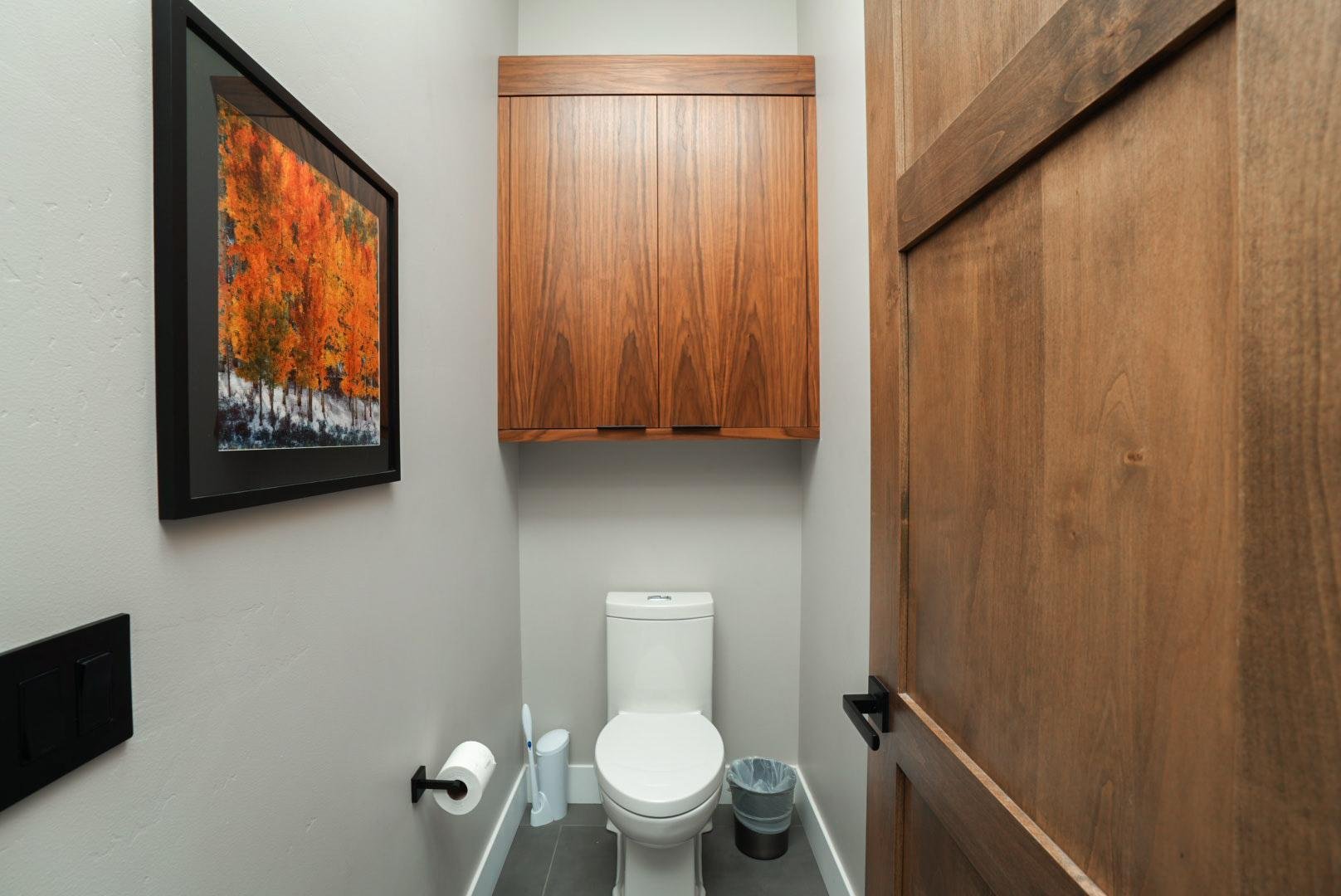 Small bathroom with a white toilet, wooden cabinet above, and a framed autumn tree painting on the wall.