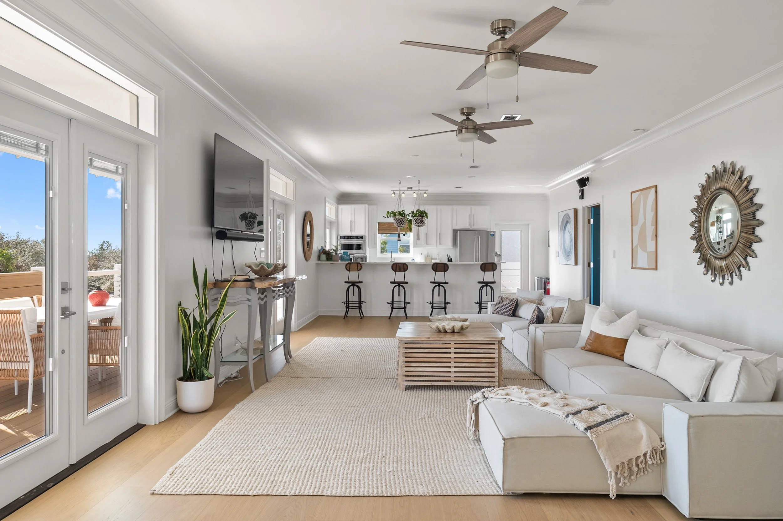 Bright open living room with white walls, hardwood floors, a beige sectional sofa with throw pillows, a wooden coffee table, and a large flat-screen TV on the wall. The space includes a kitchen area with bar stools, hanging planters, and a dining are