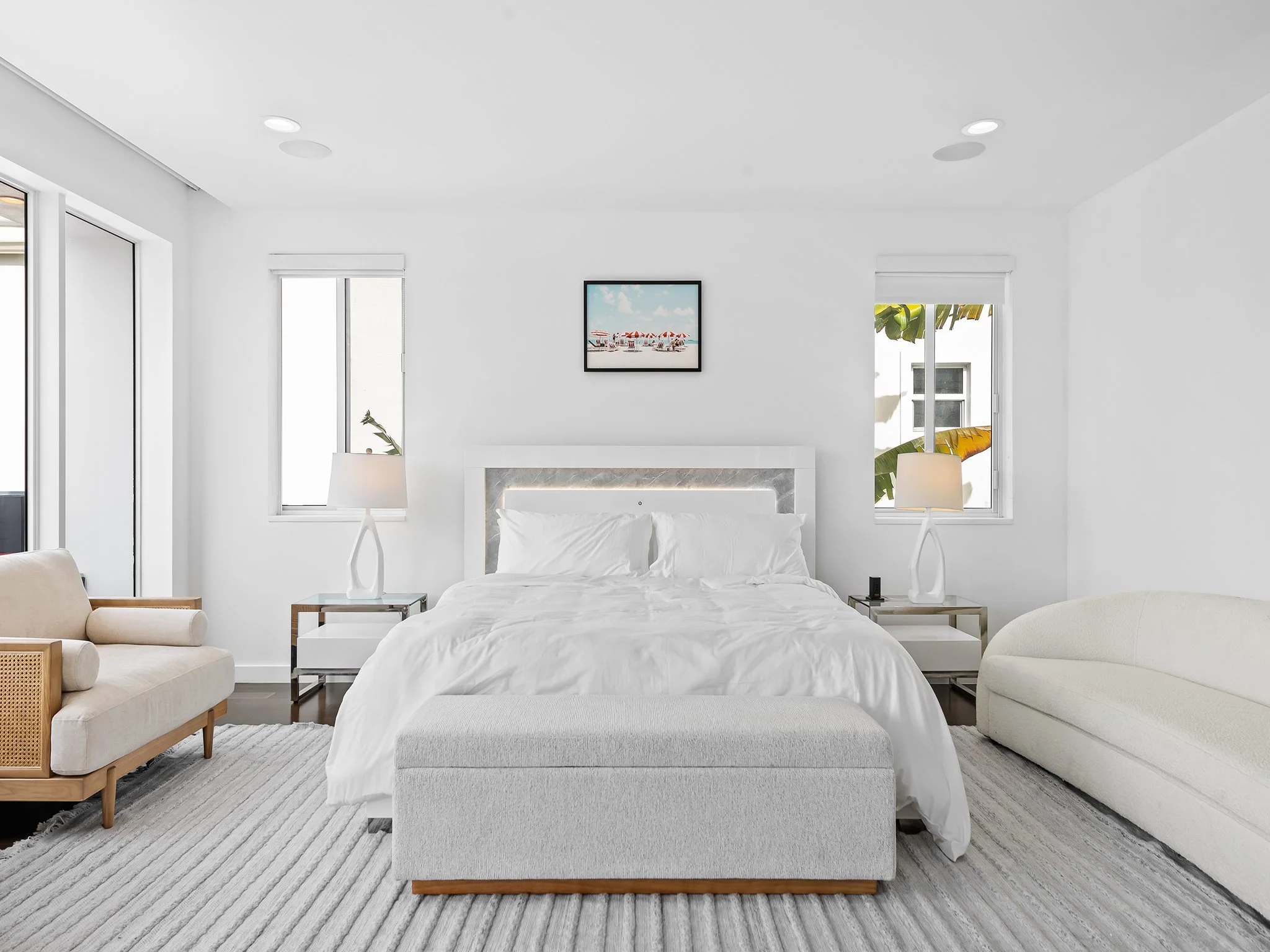 A modern, minimalist bedroom with a white bed, two side tables with lamps, and large windows letting in natural light. There's a framed beach scene hanging above the bed, and a beige sofa on the left side of the room.